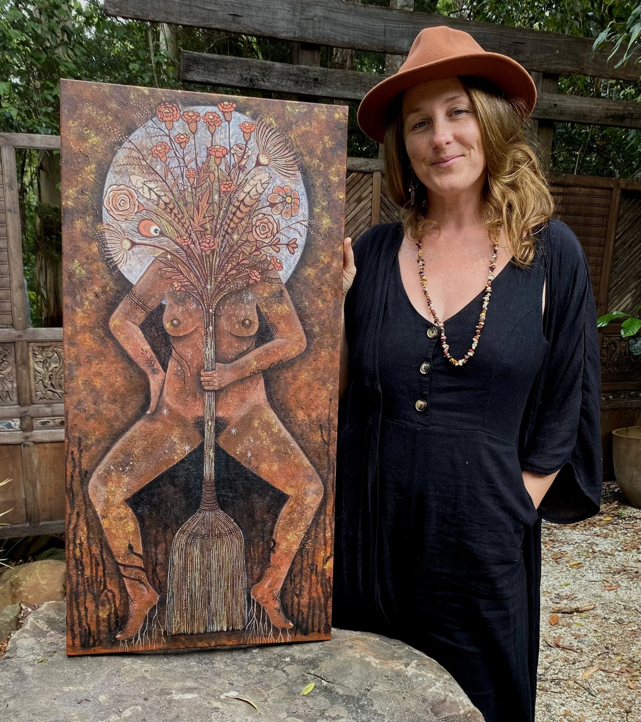 A woman wearing a brown hat and black dress standing outdoors next to a large painting of a naked woman with a bouquet of flowers and plants as her head, holding a broom, in a garden setting.