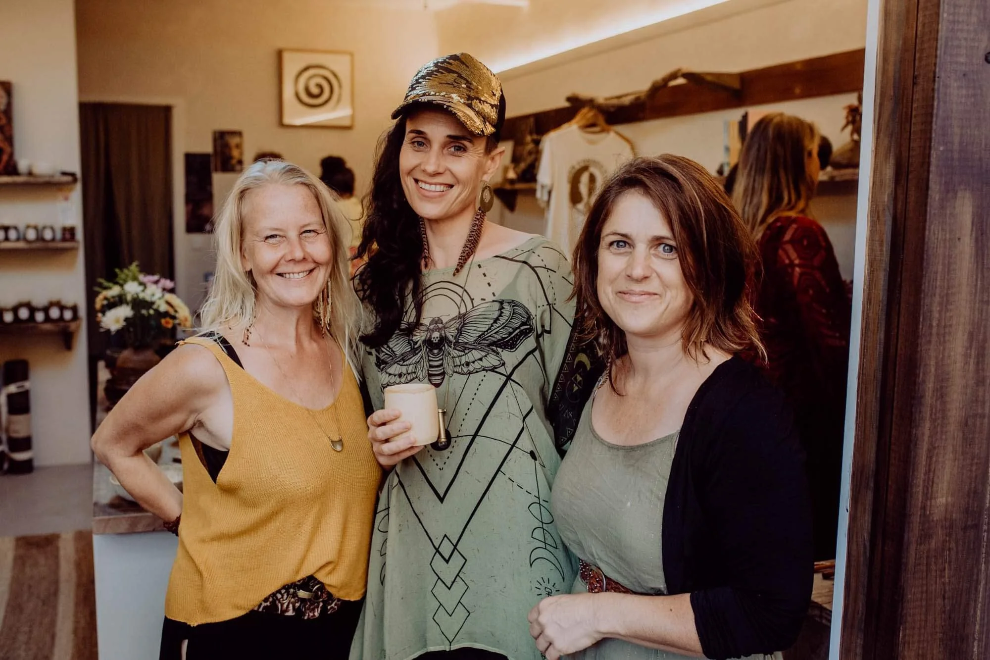 Three women smiling together indoors, one holding a cup, with shelves and artwork in the background.