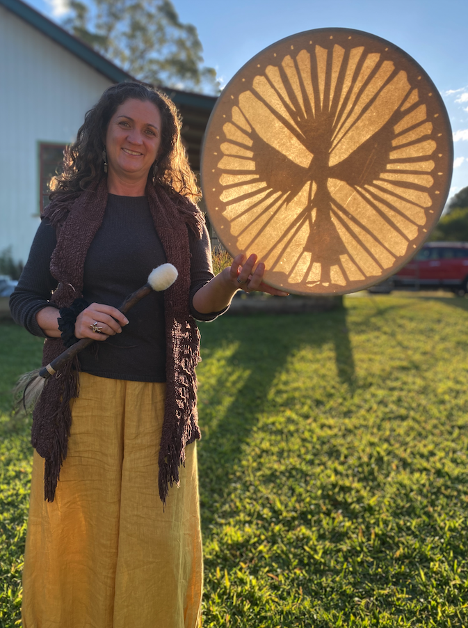 A woman standing outdoors holds a handmade drum with a silhouette of a bird on a branch surrounded by radial lines. The woman is smiling, wearing a dark sweater, yellow pants, and a brown scarf, with the sun casting a shadow behind her.