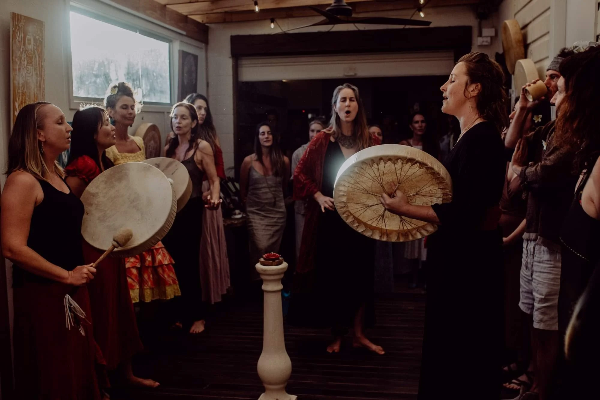 A woman in black is leading a drumming circle with a group of women gathered around her. Several women are holding drums, some with beads and other decorative items. The setting appears to be indoors with wooden walls and ceiling, and there is a wind