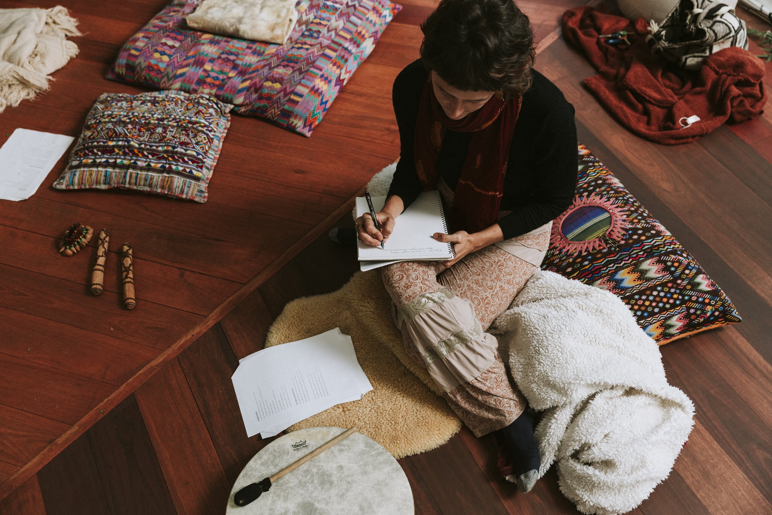 A woman sitting on a carpeted floor, taking notes on a notepad, surrounded by colorful patterned pillows, papers, and small musical instruments, with a wooden floor.