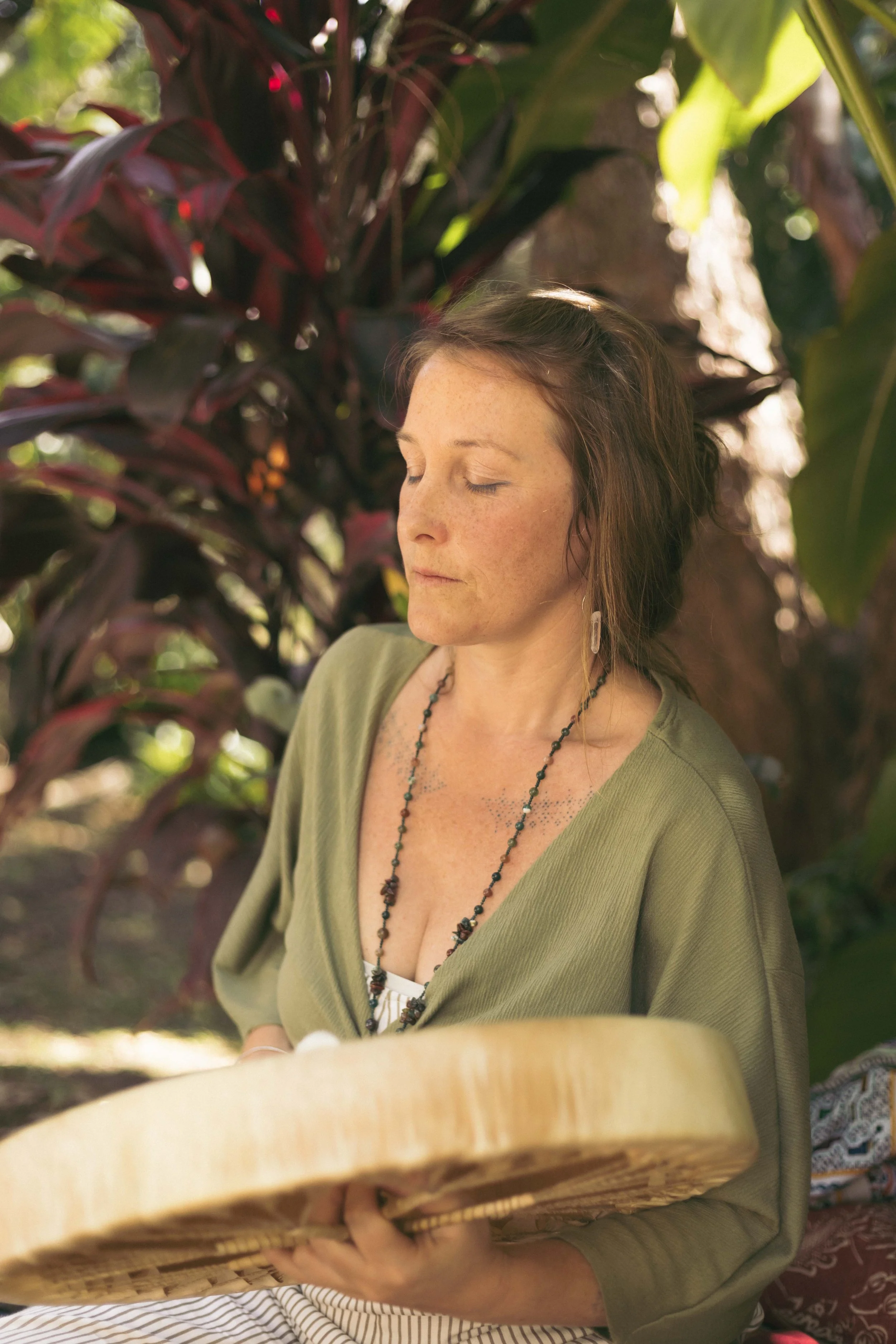 A woman with freckles and brown hair sitting outdoors, eyes closed, holding a drum and drum beater, surrounded by lush green and red foliage.