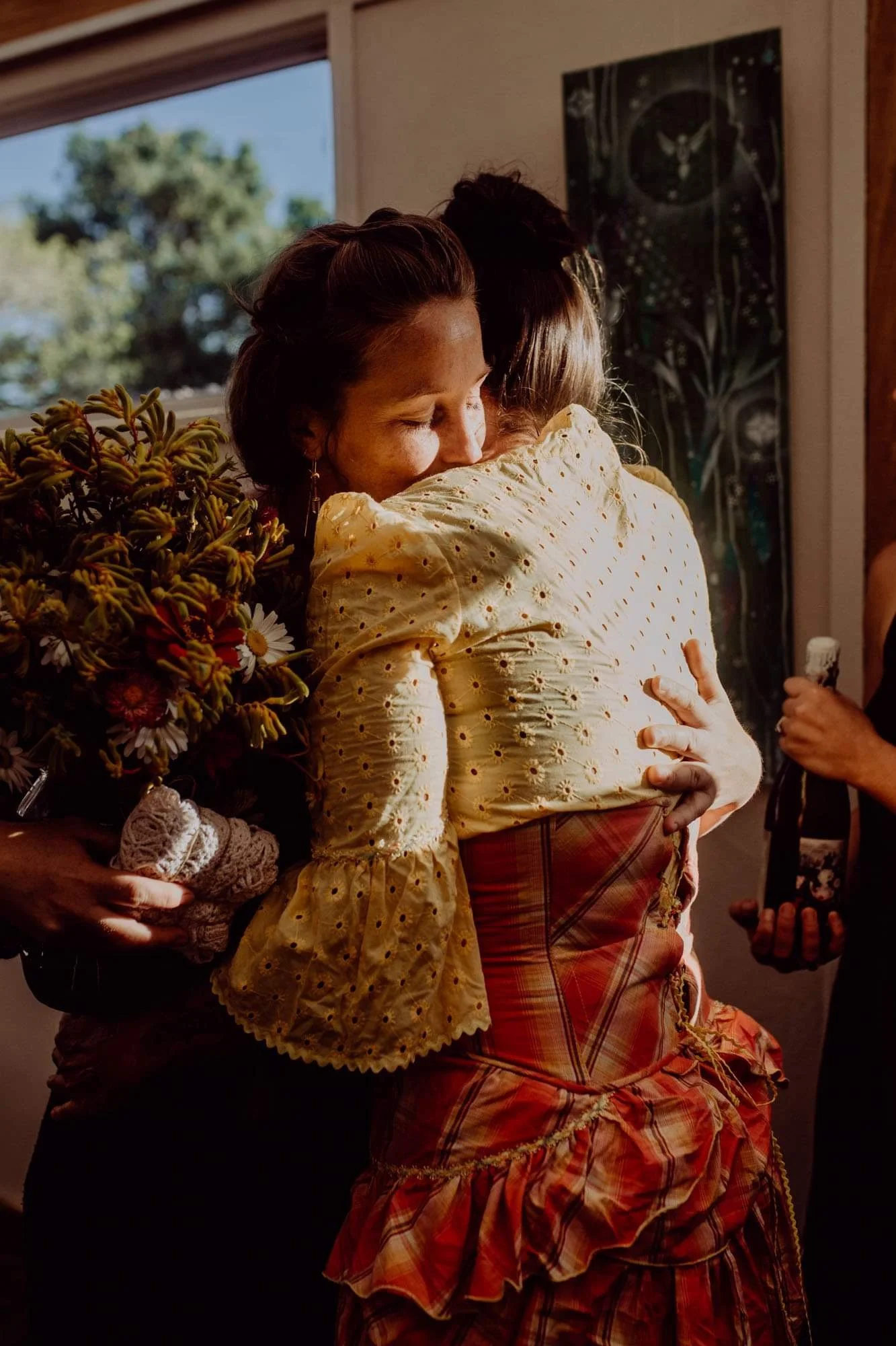 Two women are embracing, with one holding a large bouquet of flowers. The woman in the foreground has short hair, wearing a yellow eyelet blouse with puffy sleeves, and a plaid skirt. The atmosphere appears warm and emotional, possibly a farewell or 