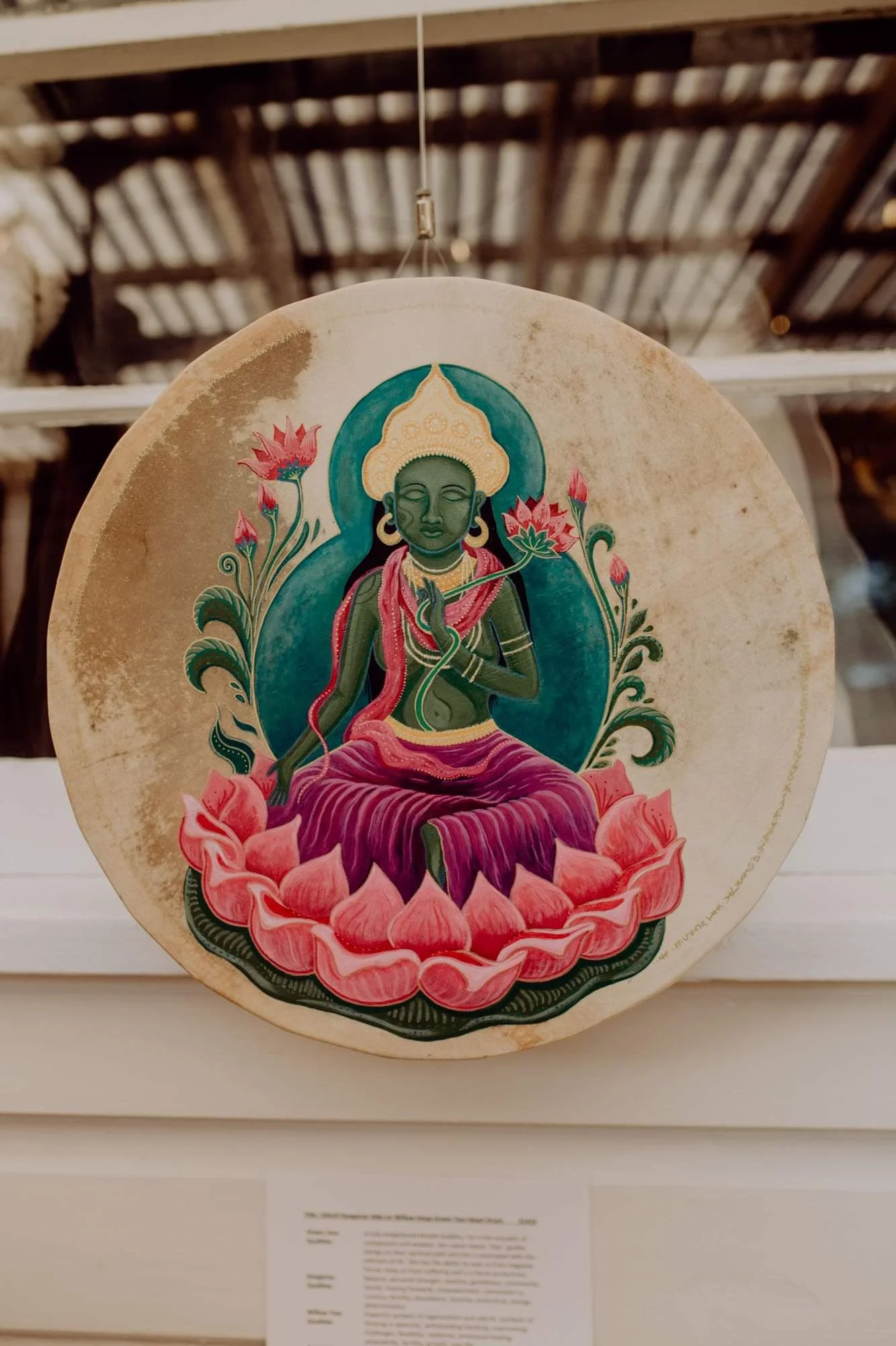 A round religious painting of a sitting deity with dark green skin, adorned with a golden crown, hoop earrings, and pink clothing, seated on a pink lotus flower with similar highlights, surrounded by a blue-green aura and pink lotus flowers on a beig