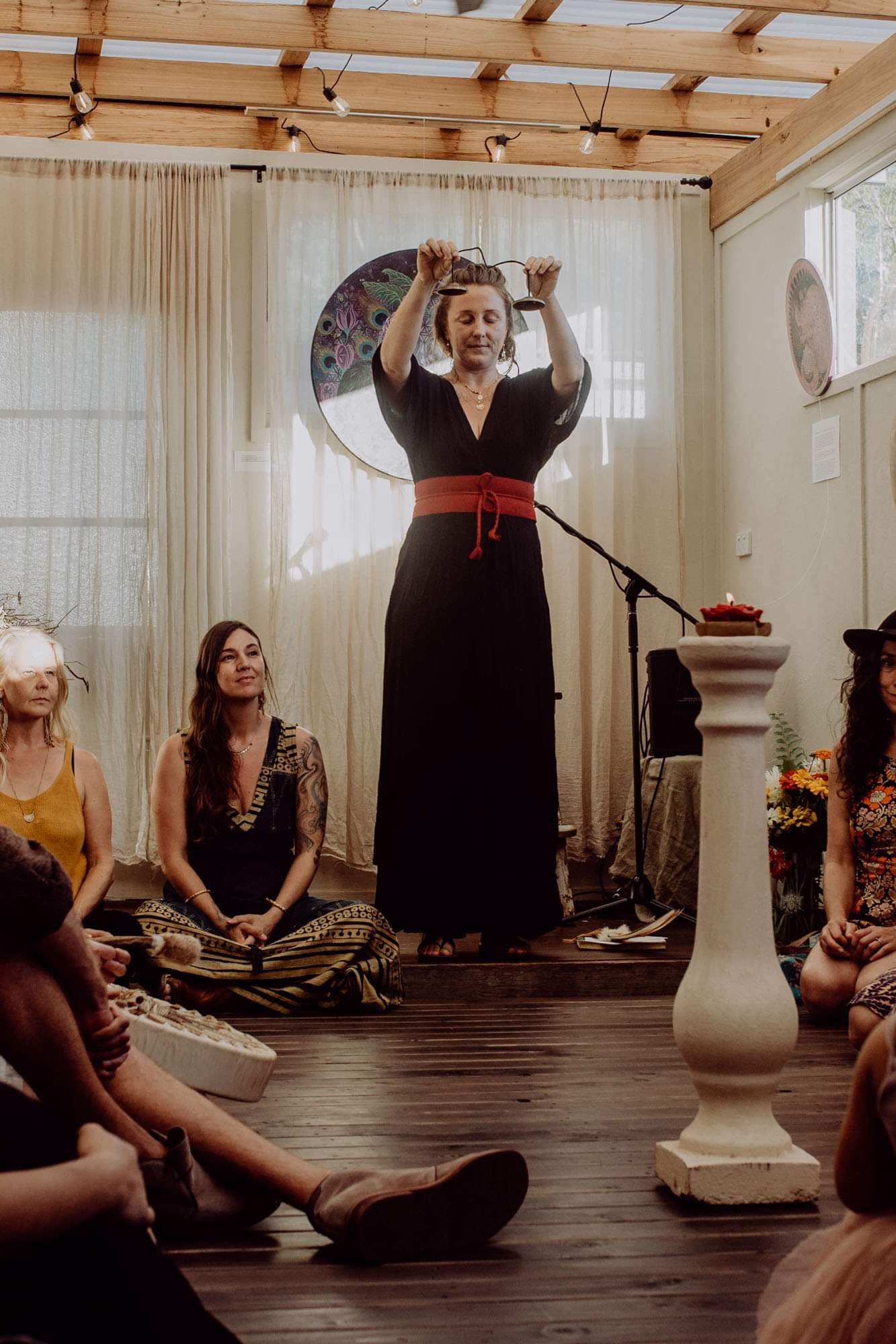 A woman standing on a stage or platform, holding an object above her head, with several other women seated on the floor around her in a cozy room with wooden ceiling beams and curtains.
