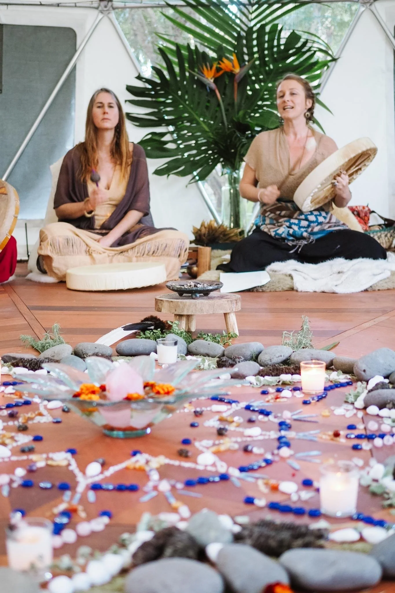 Two women sitting on the floor in a modern, brightly lit space, surrounded by lush tropical plants and a colorful arrangement of stones, candles, and decorations on the ground.