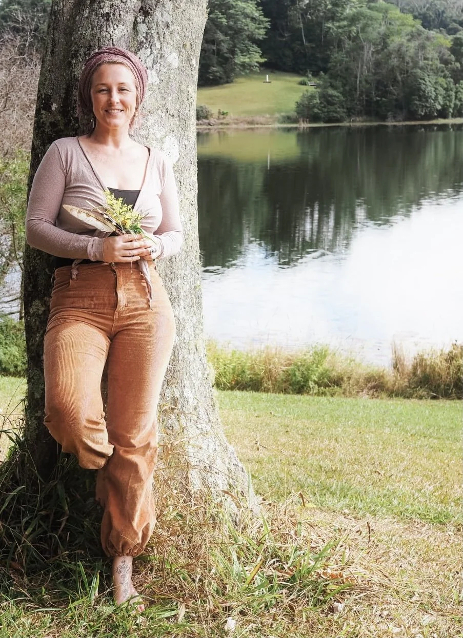 A woman stands leaning against a tree by a lake, holding a small bouquet of flowers and a fan, with green trees and grass in the background.