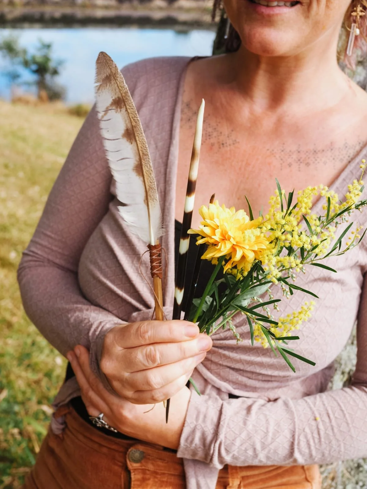 A woman in a pink textured long-sleeve top holds a bouquet of yellow flowers, a large brown feather, and a black and white striped feather, standing outdoors near a body of water.