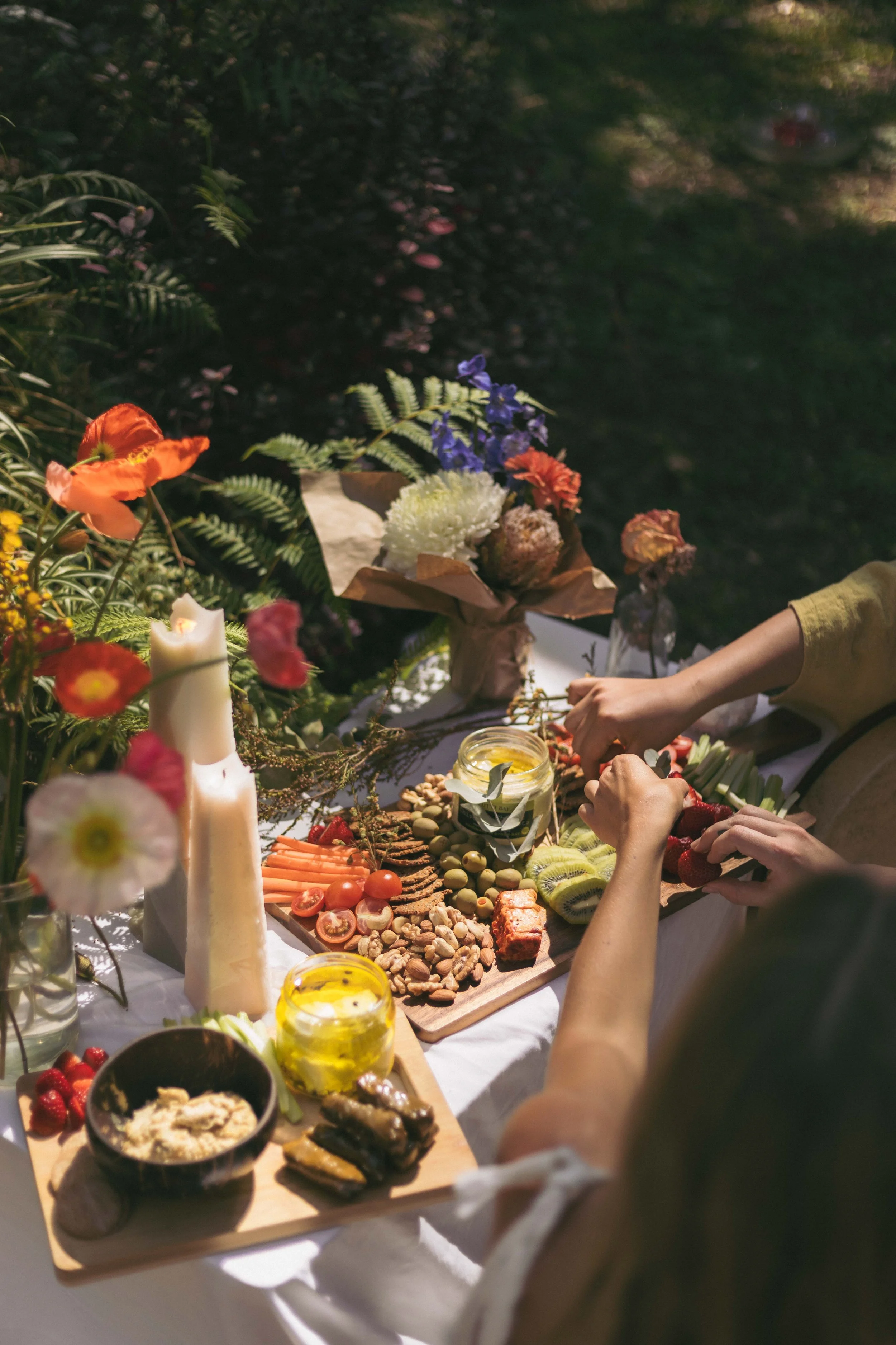 A picnic table outdoors with various snacks, fruits, flowers, and candles, surrounded by greenery.