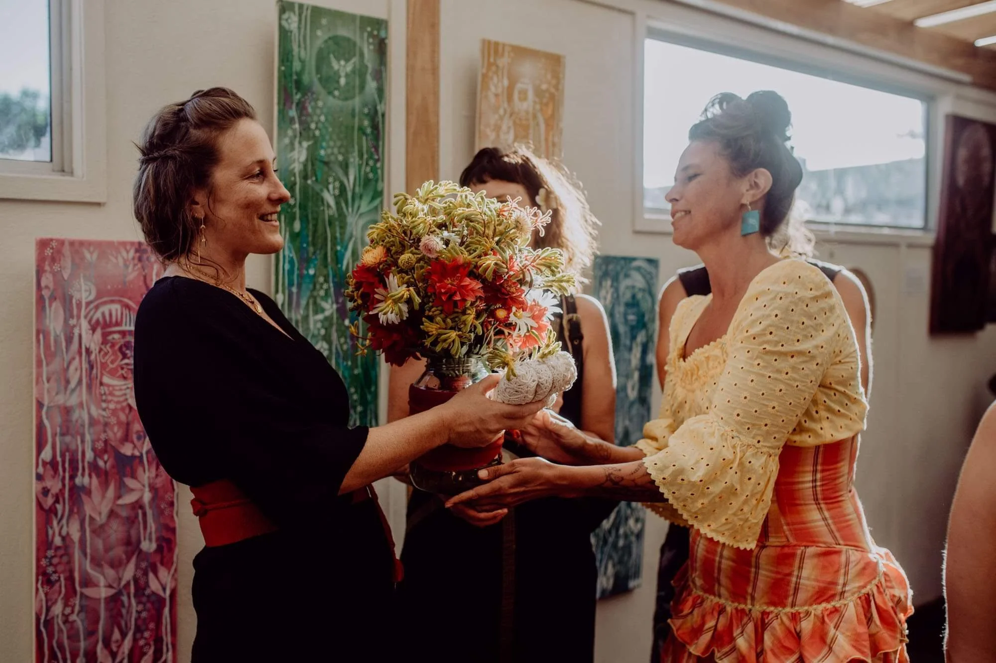 Two women exchanging a bouquet of colorful flowers in a well-lit art gallery with paintings on the wall.