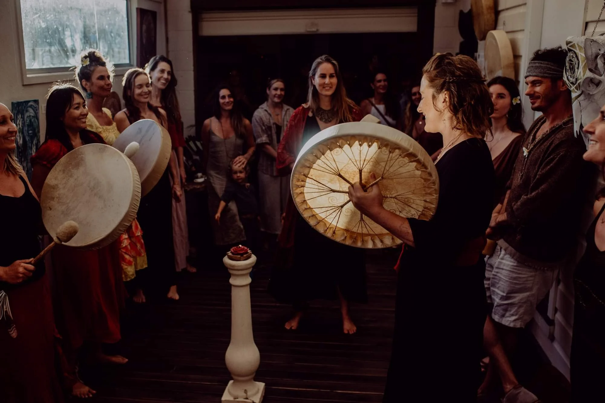 Group of people participating in a drum circle indoors, with some holding traditional hand drums and one woman holding a large umbrella-like object, in a cozy wooden room with a firewood stack and window.