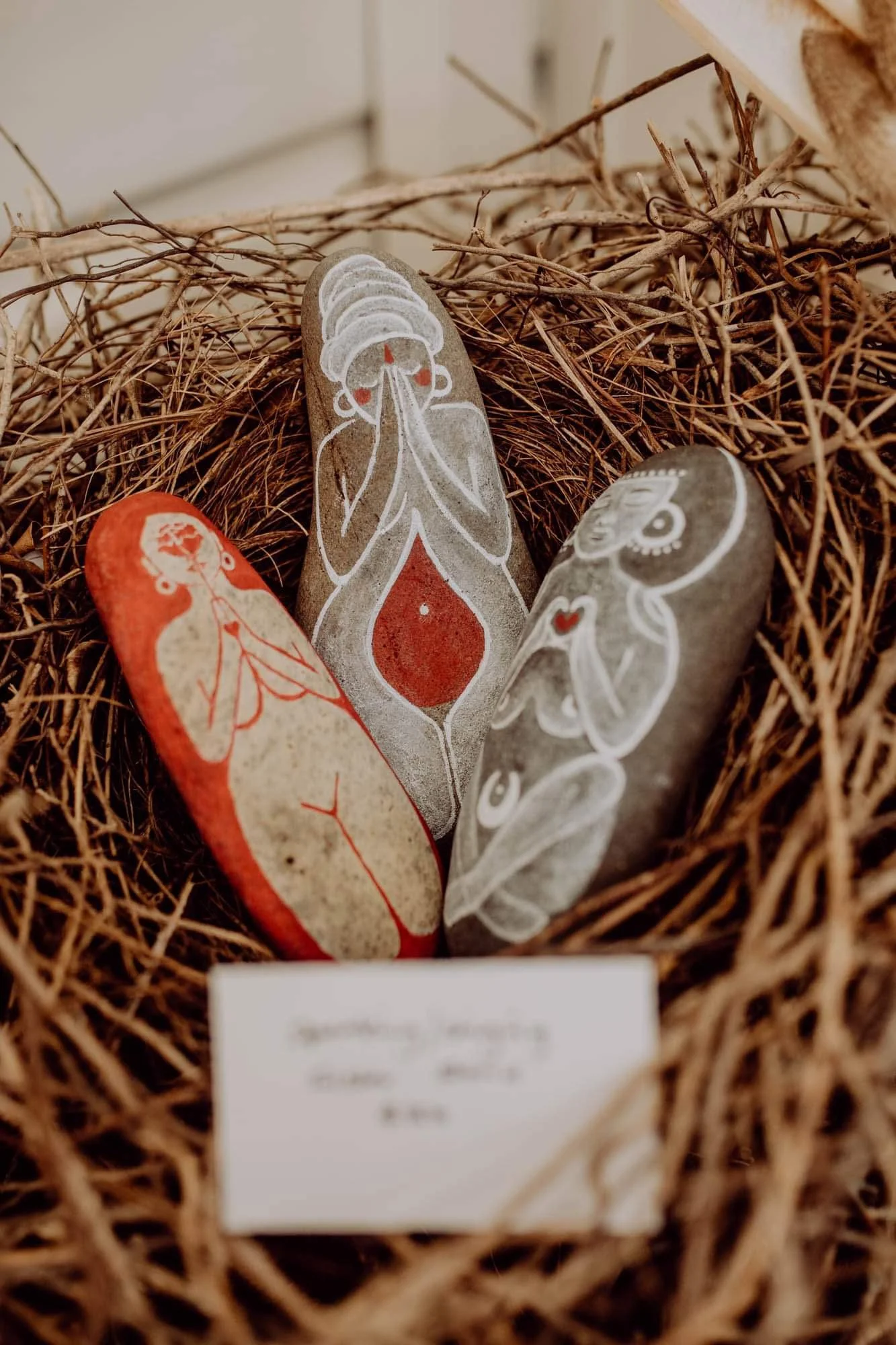 Three painted stones featuring abstract human figures, arranged in a nest of twigs, with a blurry white tag in the foreground.