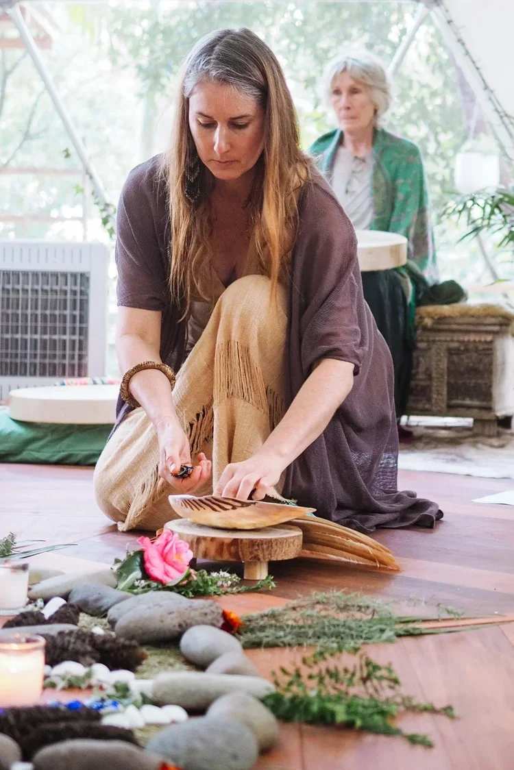 A woman with long hair, dressed in earthy tones, kneeling on the floor and arranging objects on a small wooden table during a spiritual or healing ceremony. In the background, an older woman with gray hair wearing a green jacket observes. The setting appears to be a bright indoor space with natural light, decorated with rocks, candles, flowers, and greenery.