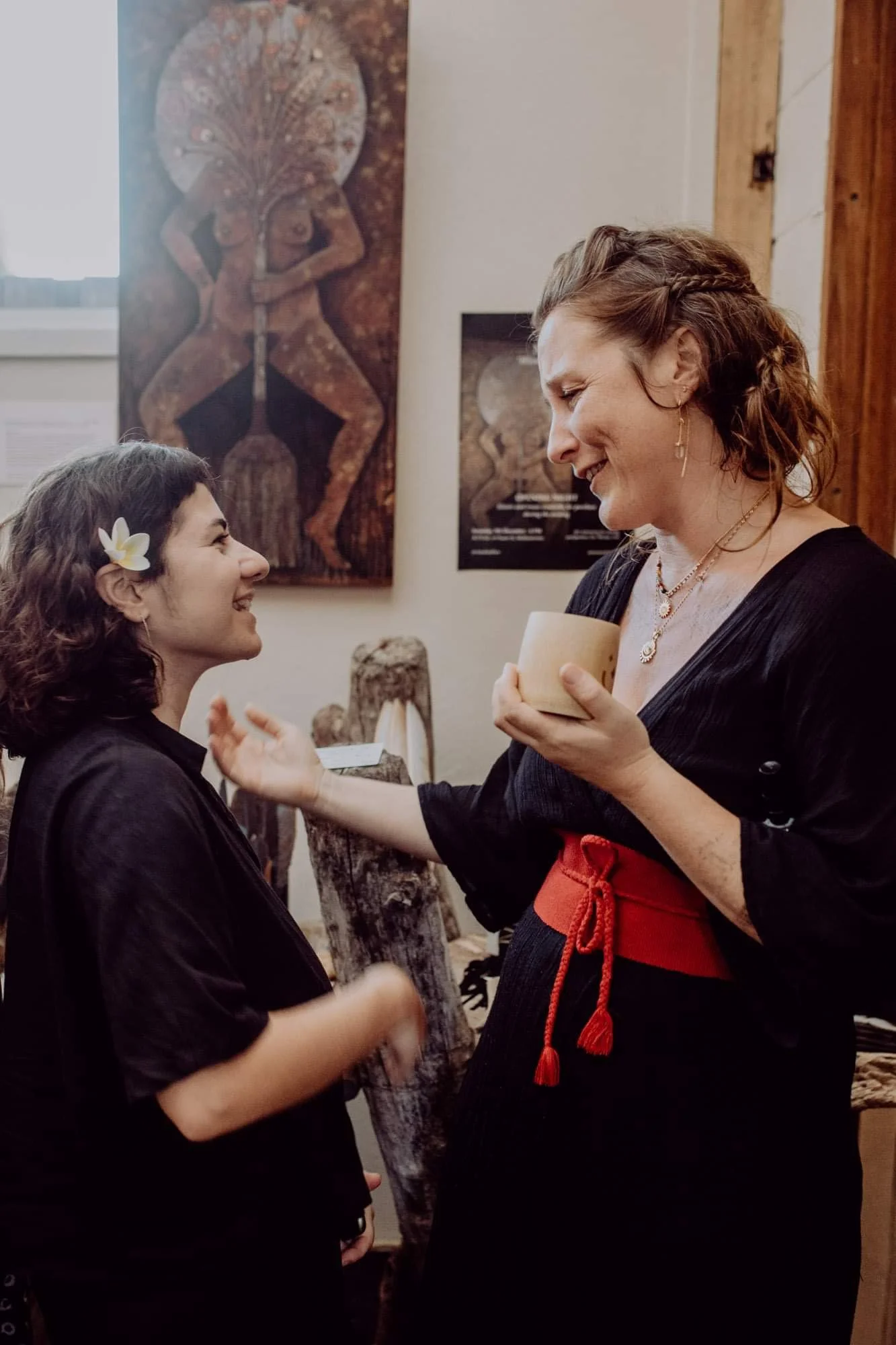 Two women smiling and engaging in conversation at an art gallery, with Aboriginal-style artwork on the wall behind them.