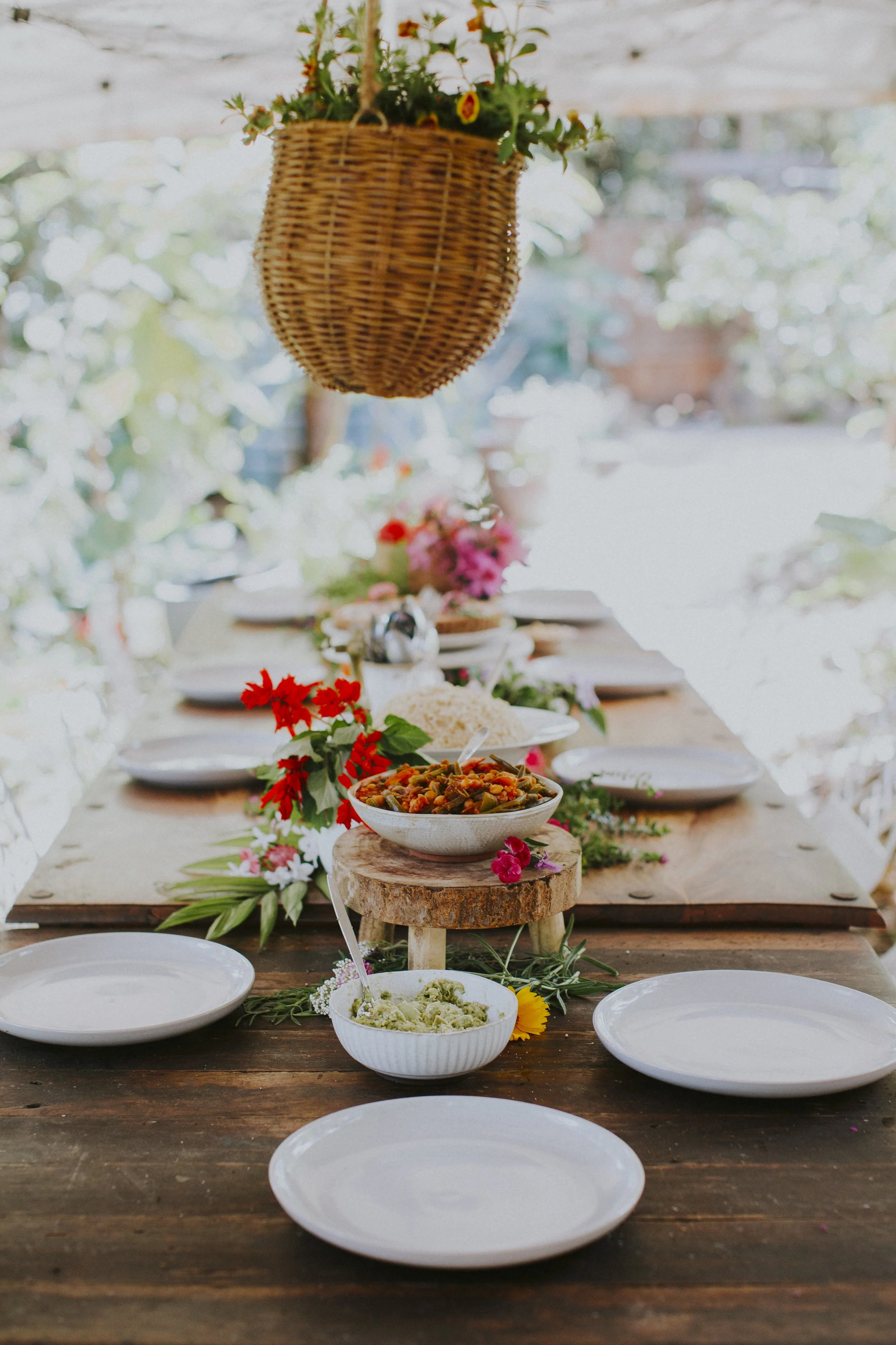 A long wooden dining table set with white plates, with a rustic centerpiece of flowers and bowls of food, decorated with a hanging wicker basket with flowers inside, outdoors with greenery in the background.
