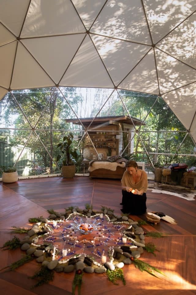 A woman sitting on a wooden dance floor inside a geodesic dome, arranging candles and decorative items on a circular arrangement of stones and greenery, with natural light filtering through the dome's panels.