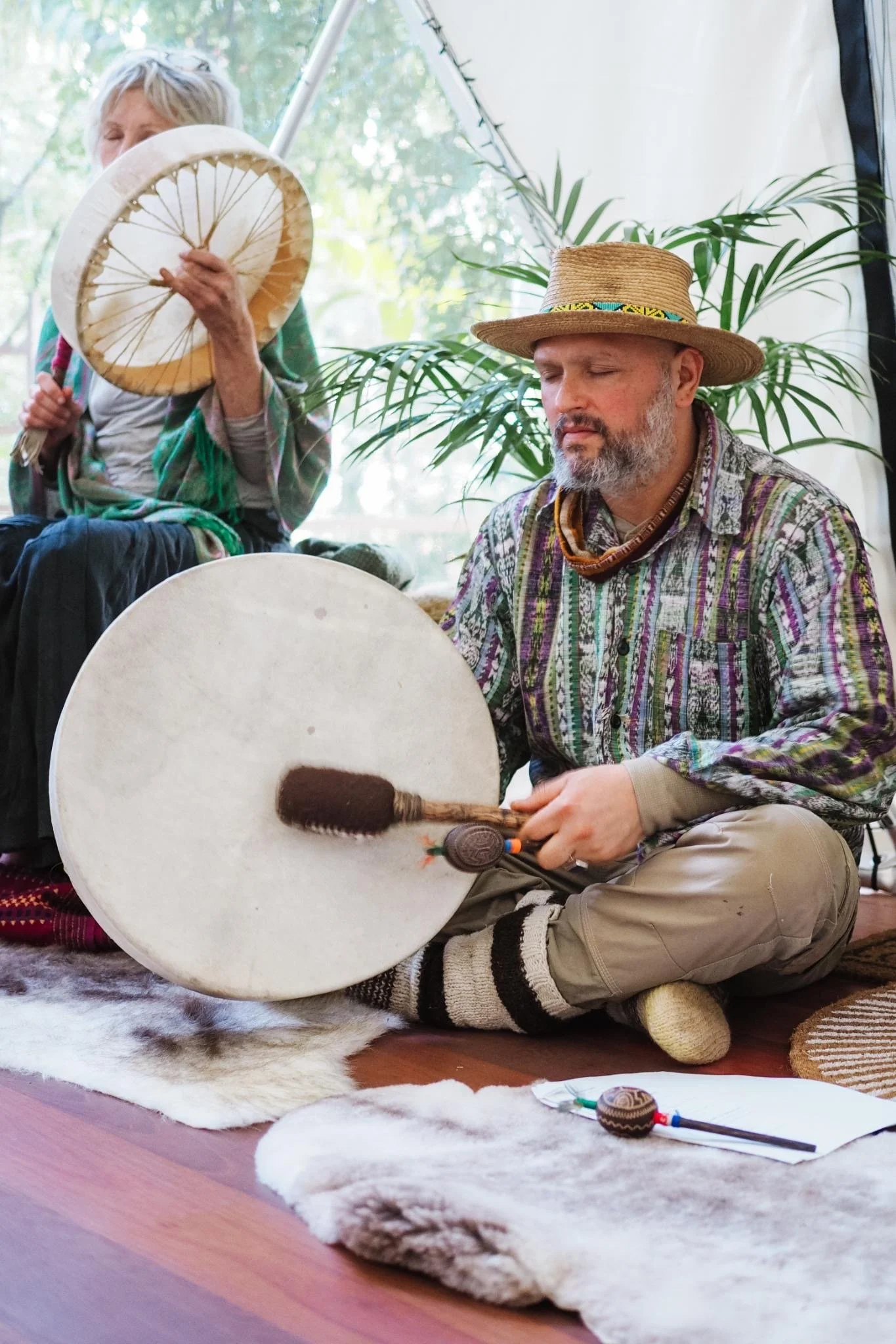 A man with a beard wearing a straw hat and a colorful patterned shirt is sitting cross-legged on a fur rug, holding a large drum and a mallet. An older woman with short gray hair is sitting nearby. The scene appears to be inside a tent with natural light, plants, and greenery in the background.
