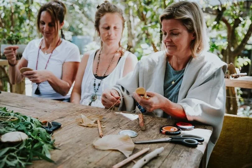 Three women sit at a wooden outdoor table surrounded by greenery, engaged in arts and crafts, working on sewing projects with fabric, scissors, and other tools.