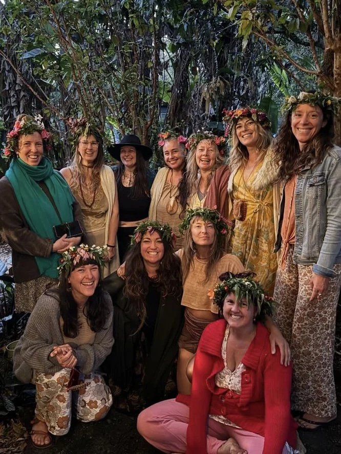 A group of women wearing flower crowns smiling together outdoors in front of a lush, green background.