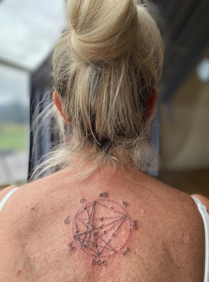 A sacred tattoo of a geometric diagram with various lines, points, and labels on a person's upper back near the shoulder.