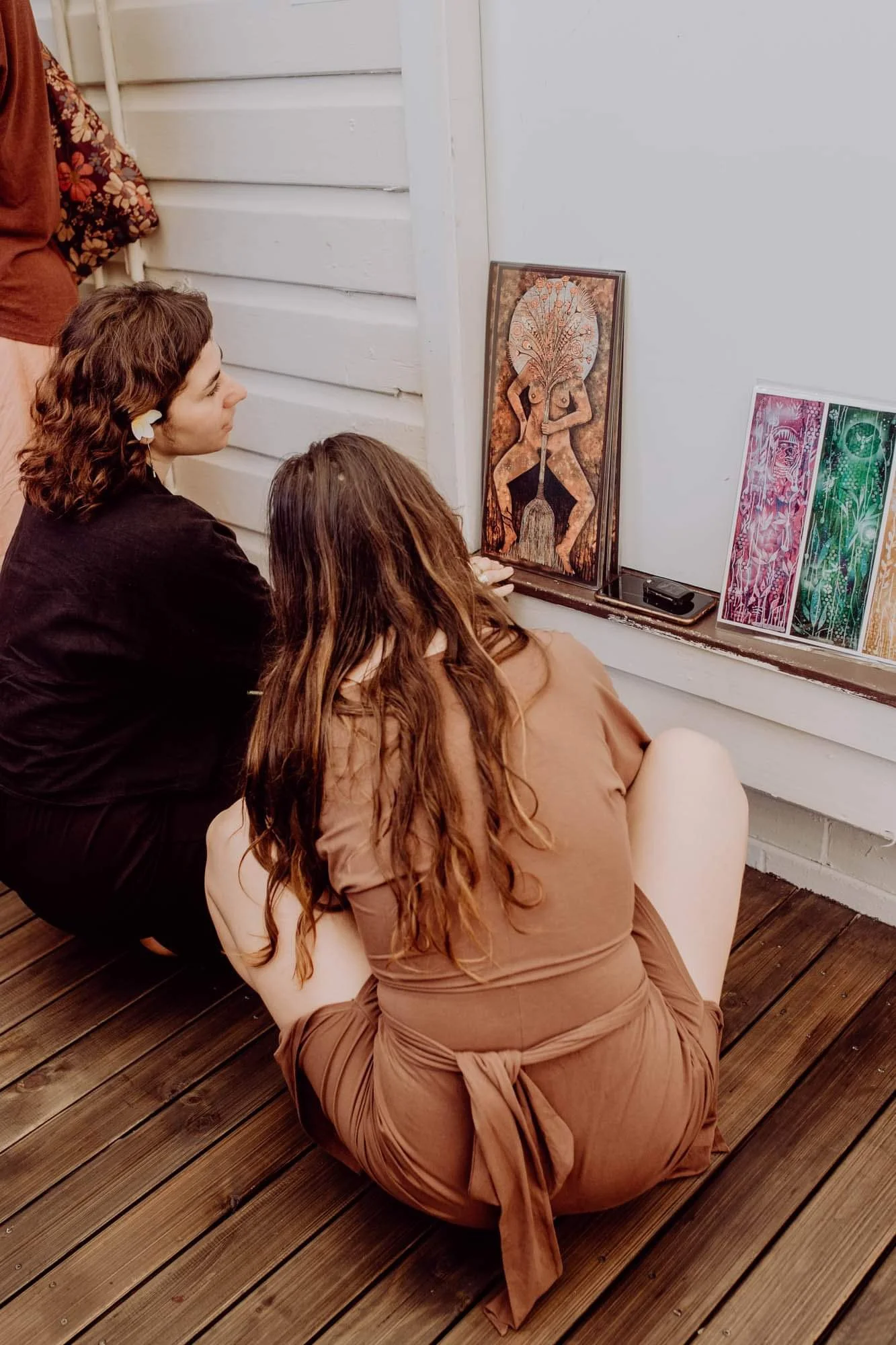 Two women, one with curly brown hair and a white flower in her hair, squatting on a wooden deck and looking at artwork on a window ledge. One is wearing a black top, the other a brown dress. There are three colorful prints and a framed piece of art d