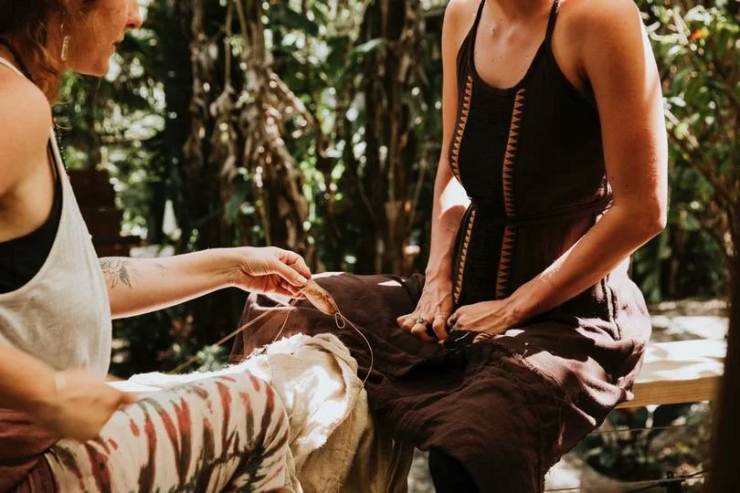 Two women sit outdoors in a lush, wooded area, engaging in a traditional craft or textile activity with fabric.