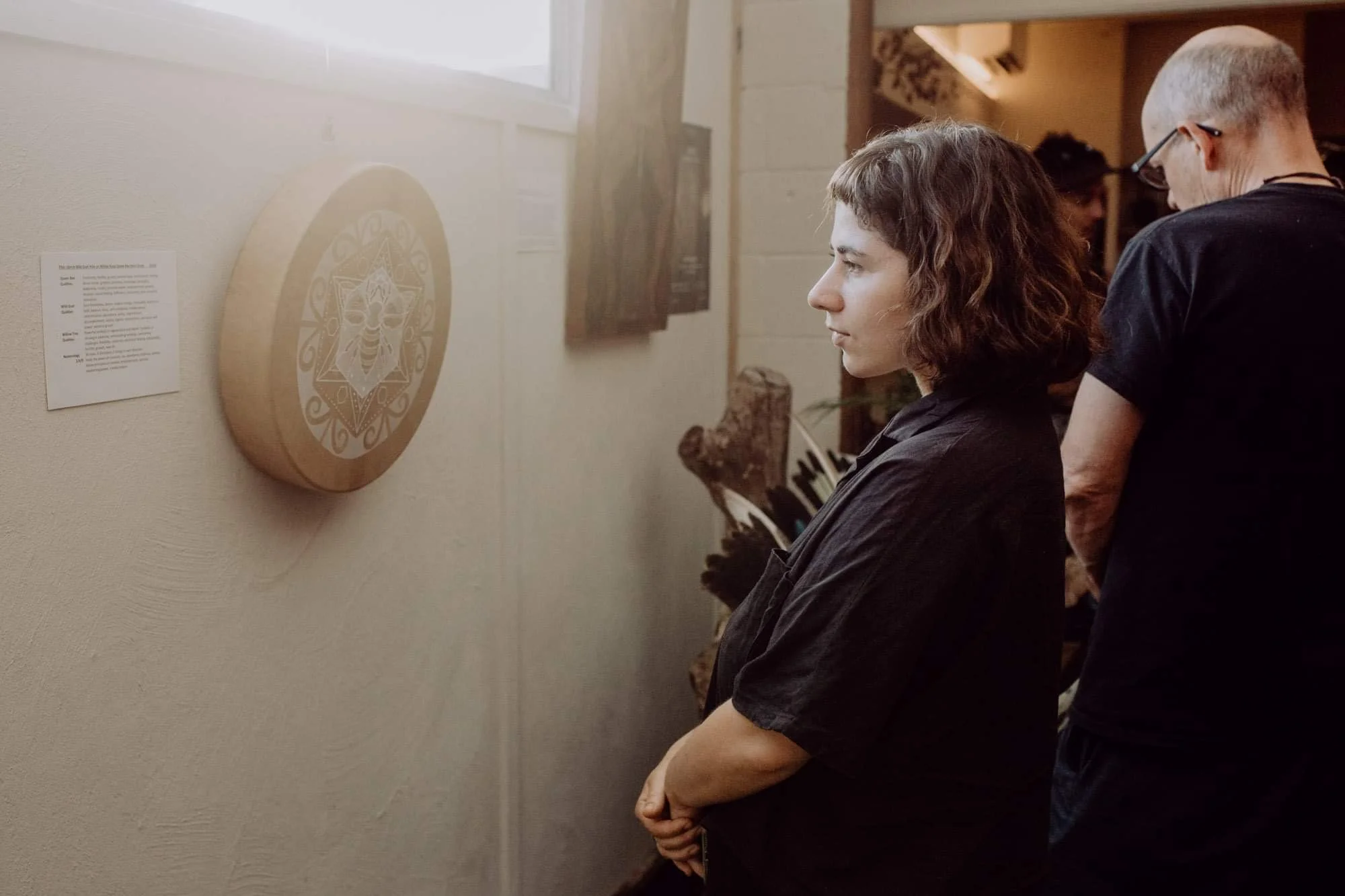 Two women are viewing art on a gallery wall; the woman in the foreground has wavy brown hair and is wearing a black shirt, her hands folded, while the other woman has short blond hair and glasses, also in a black shirt, looking at artwork.