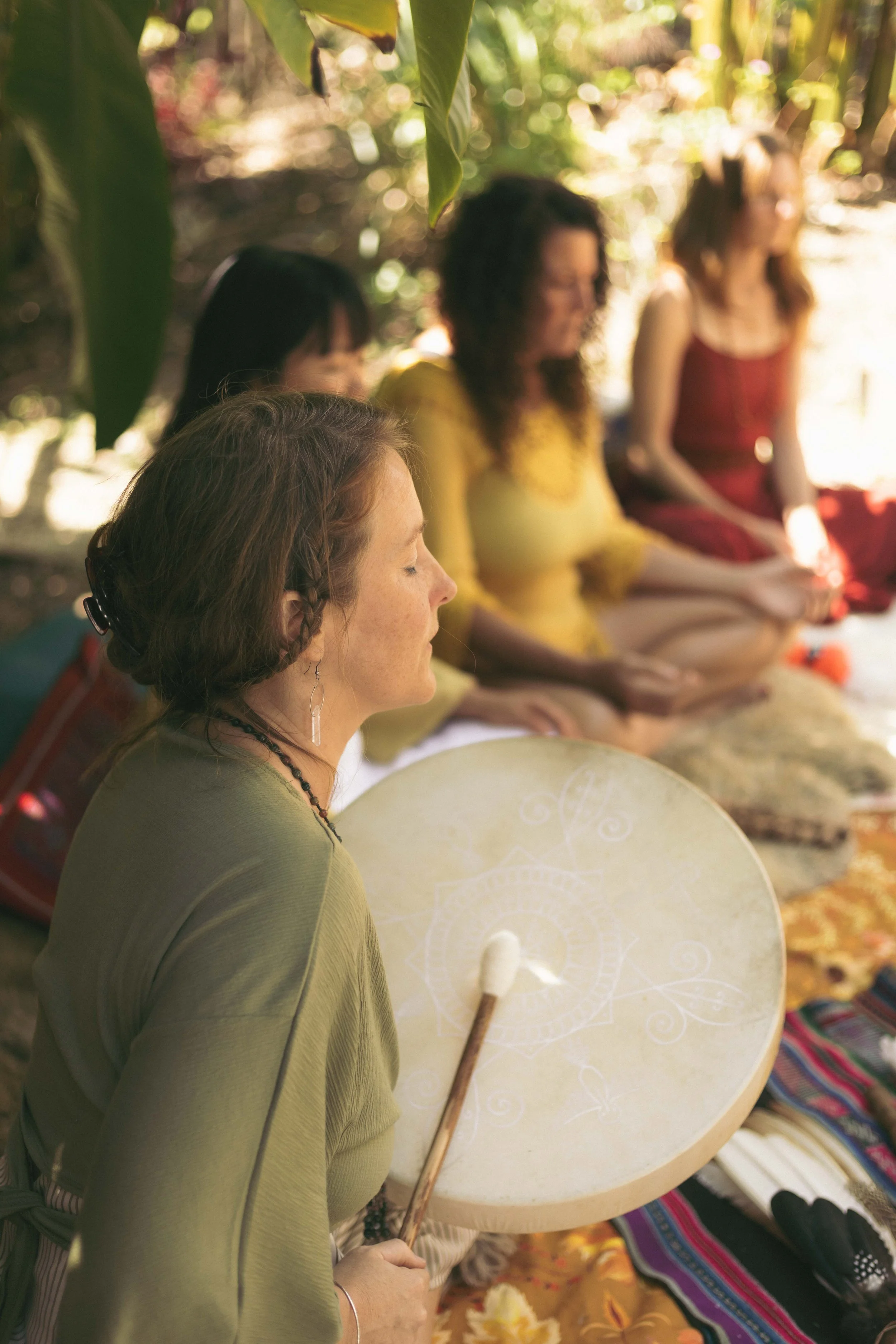 A woman in a green dress with closed eyes, holding a large drum and a drum beater, participating in a meditation or spiritual ceremony outdoors in a jungle-like setting with other women sitting in the background.