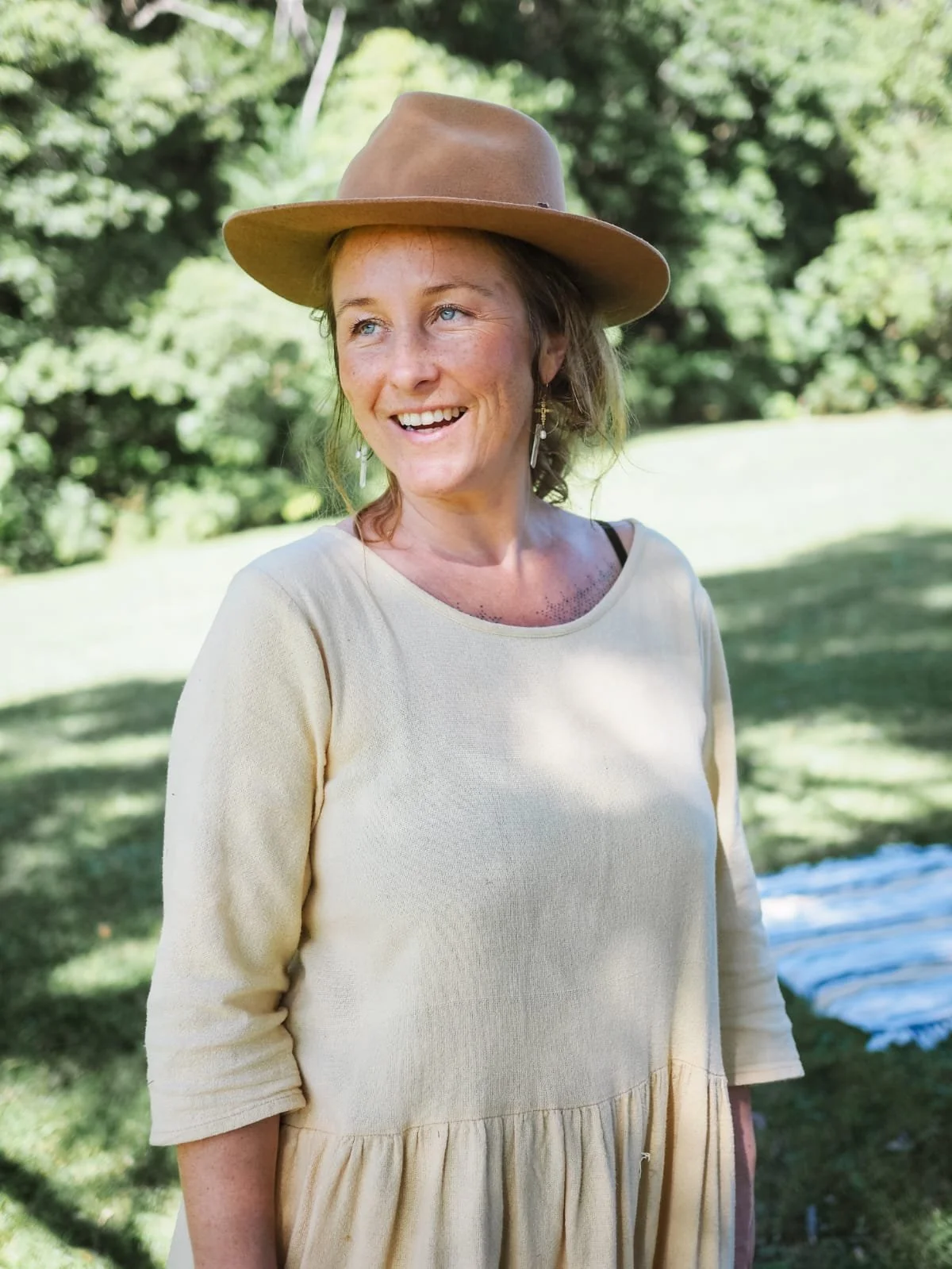 A woman with blonde hair and blue eyes smiling outdoors, wearing a beige dress, a tan wide-brimmed hat, and earrings, with greenery in the background.