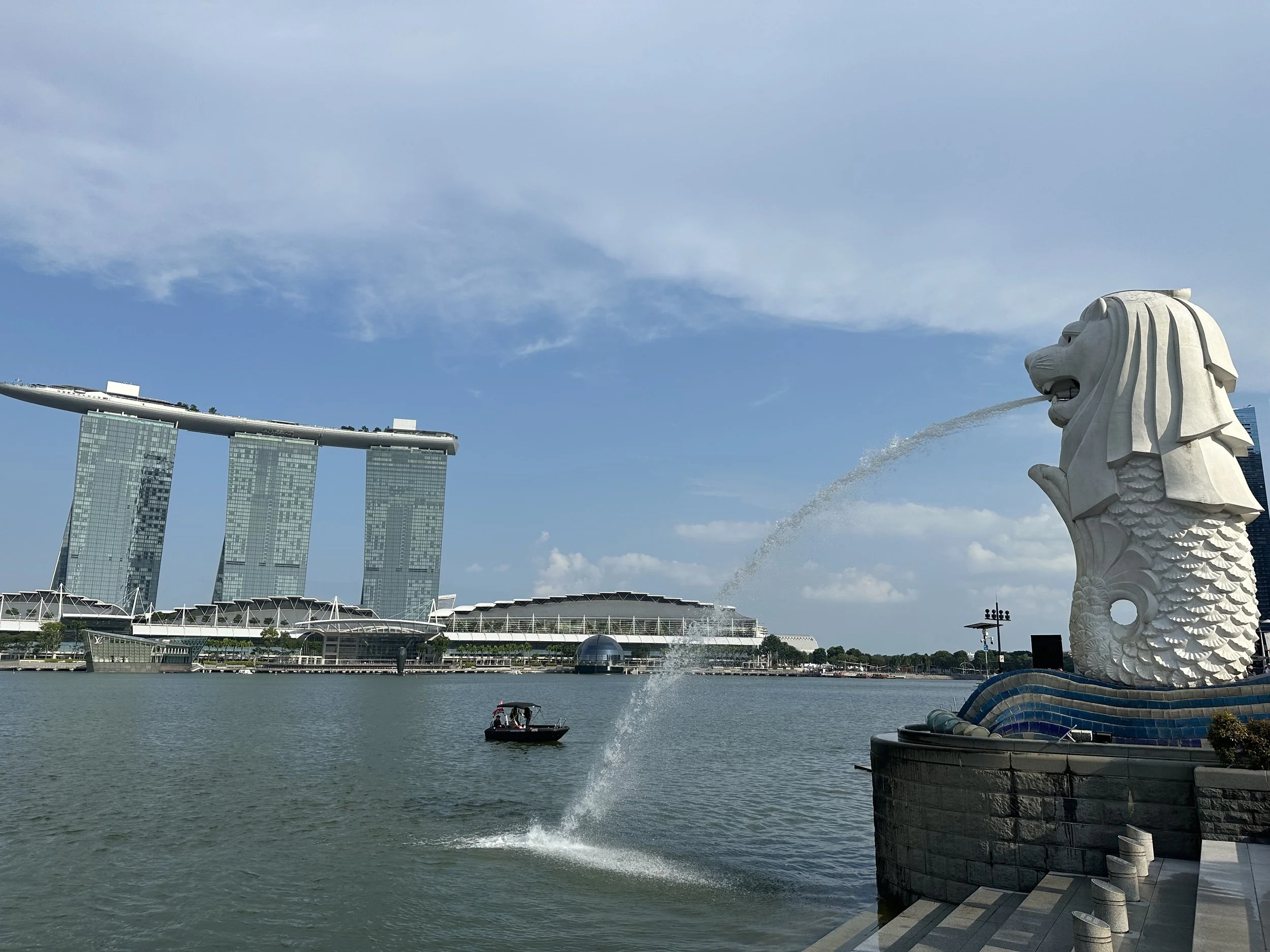 A view of the Marina Bay Sands hotel with three towers and a boat in the water. The Merlion statue spouting water is in the foreground on the right side.