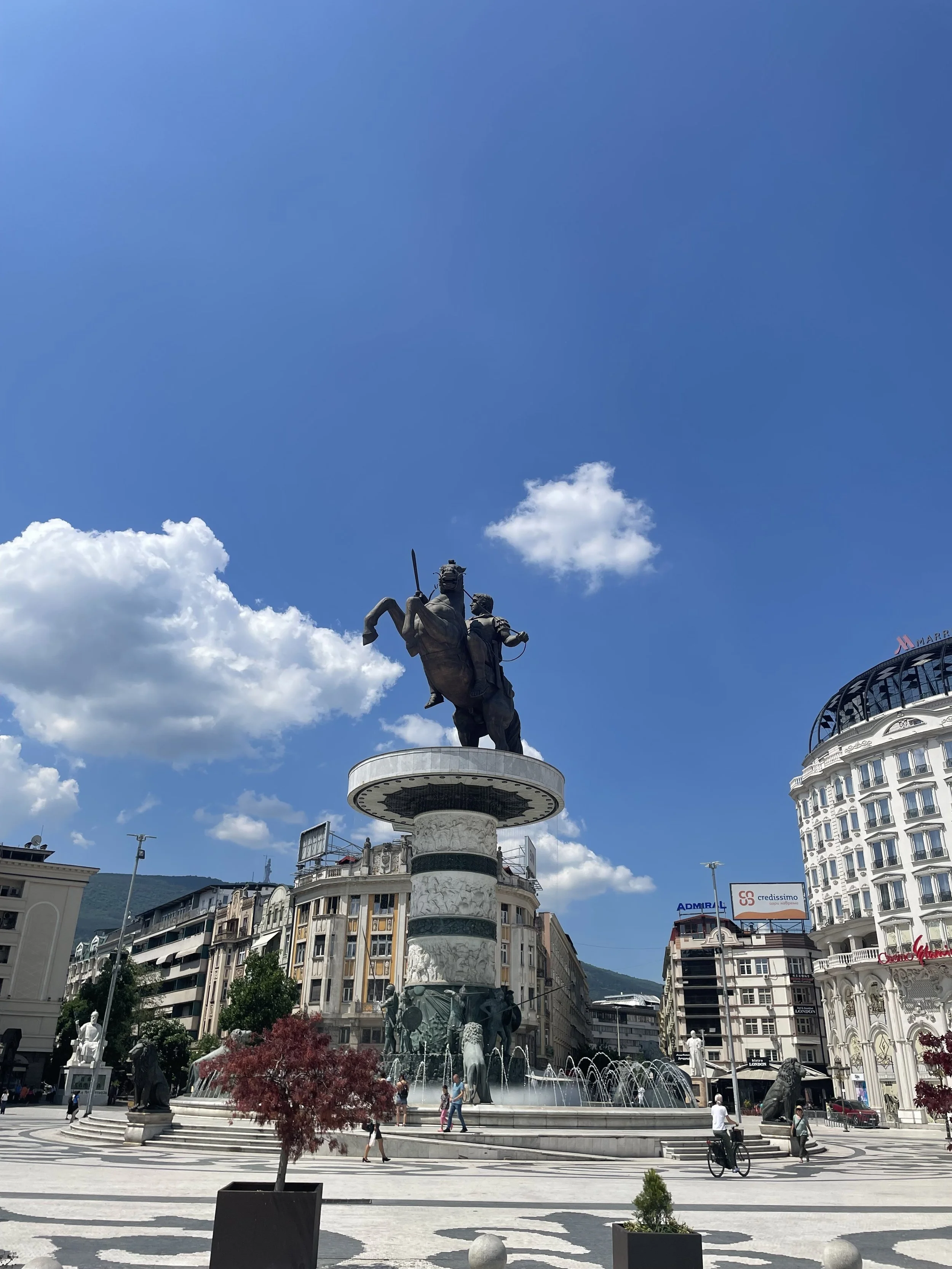 Statue of two men on horseback with a fountain and city buildings in the background under a blue sky with clouds.
