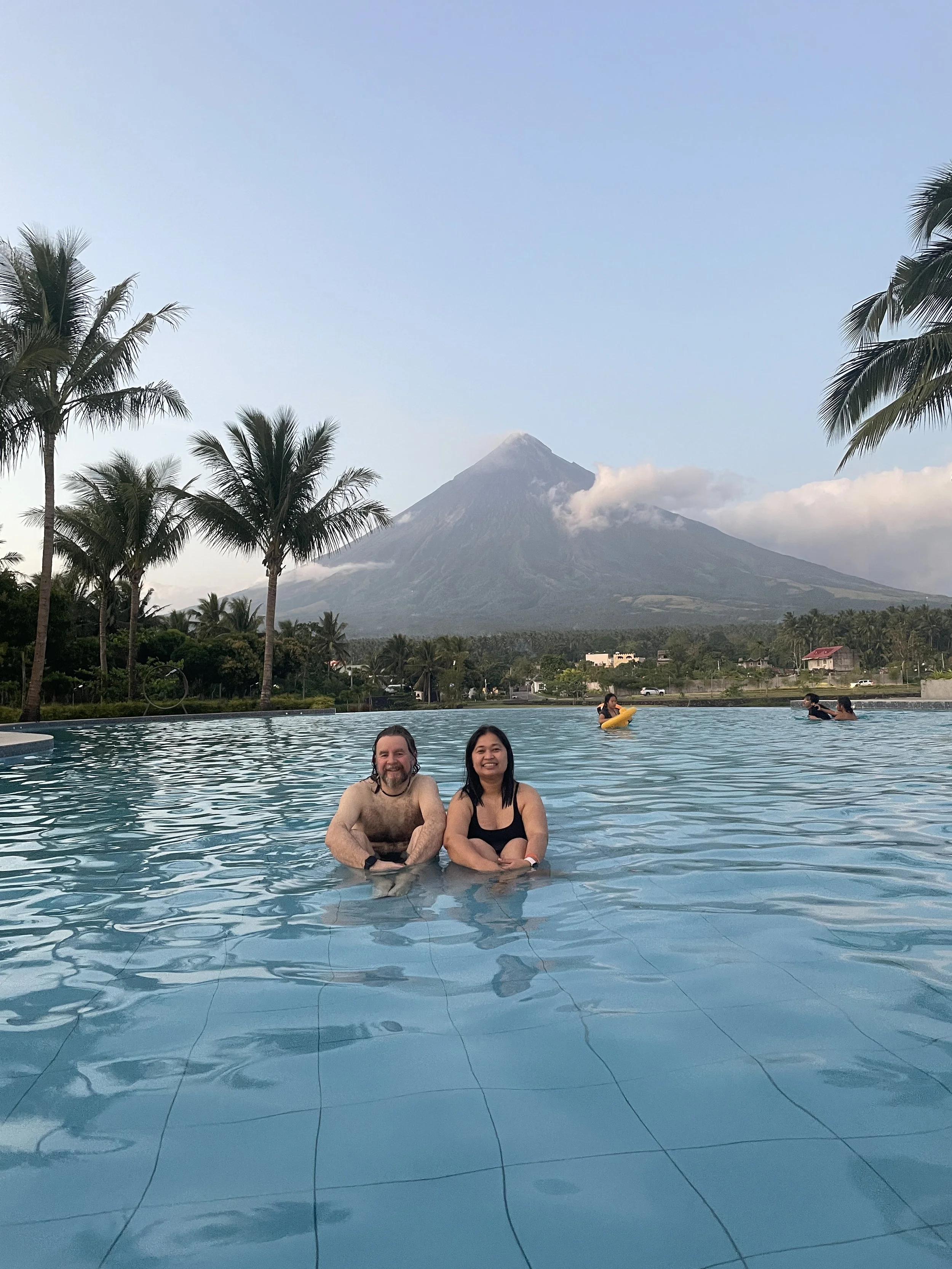 Two people smiling in a swimming pool with a mountain and palm trees in the background.