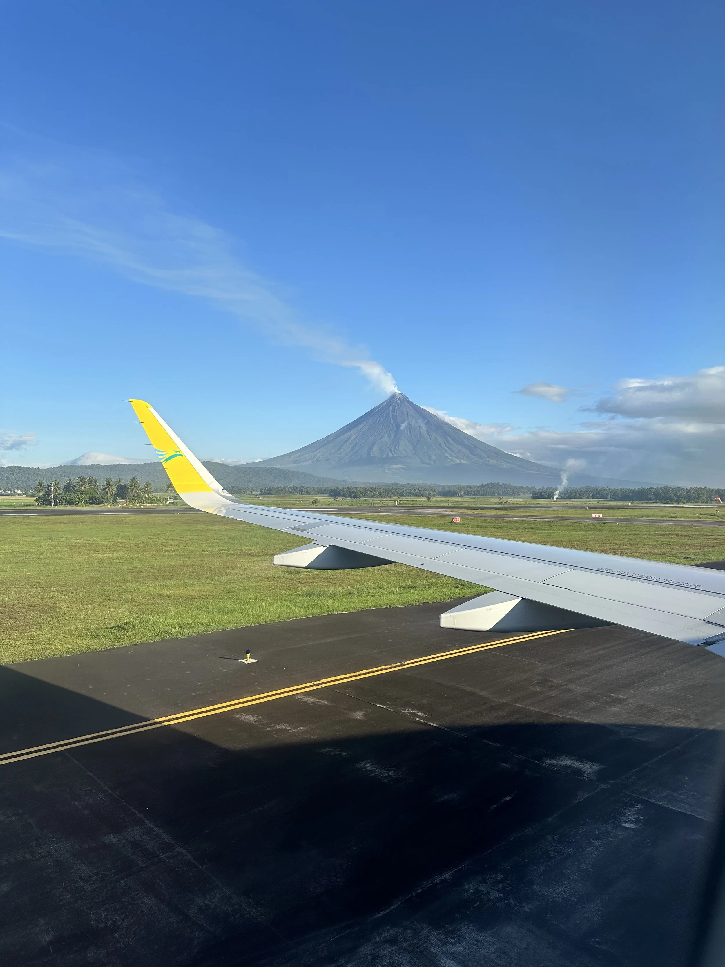 View from airplane window showing airplane wing on runway with a volcano erupting in the background under a blue sky.