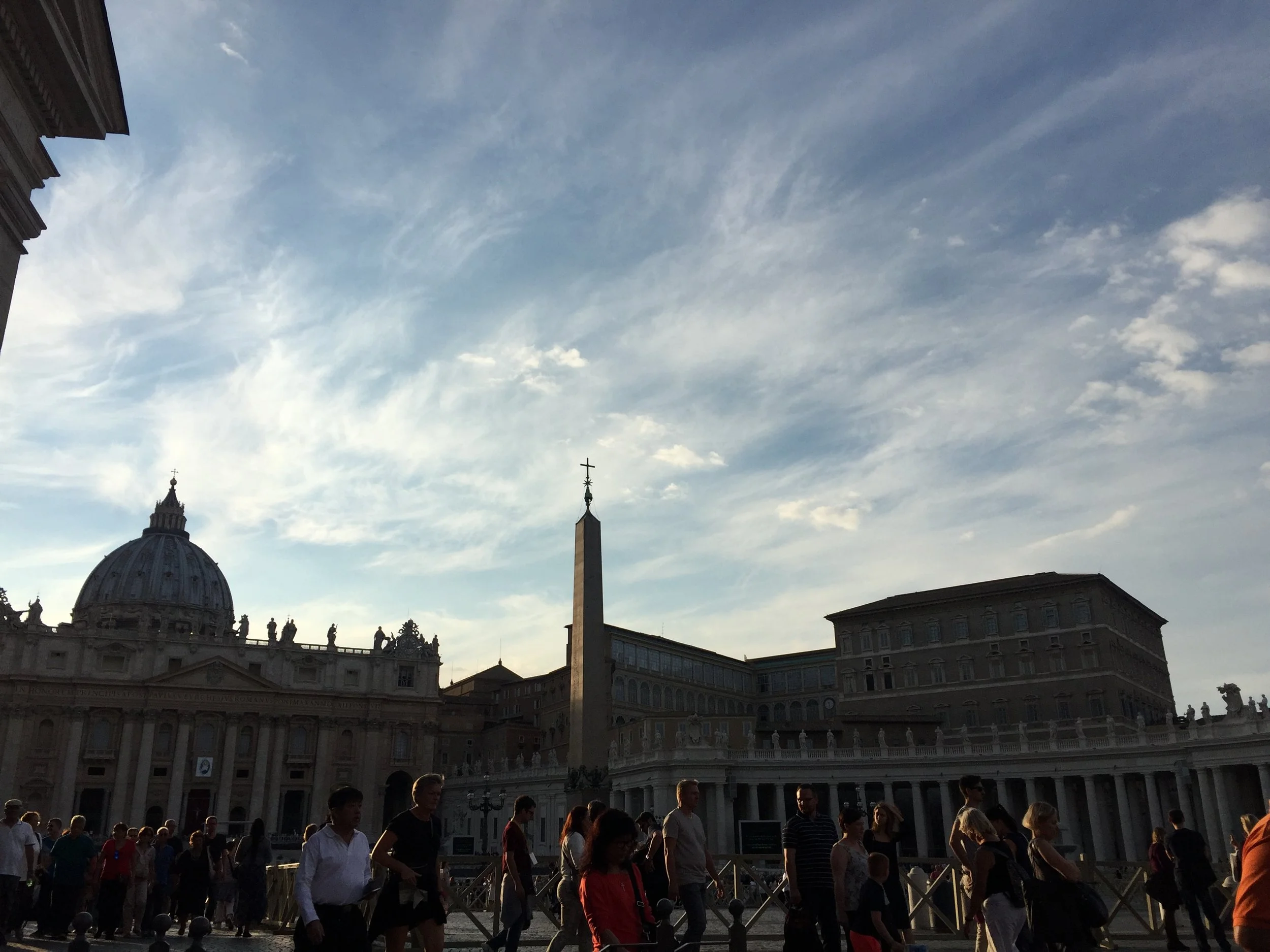 People gather outside St. Peter's Basilica in Vatican City, with the obelisk and historic buildings visible, under a partly cloudy sky.