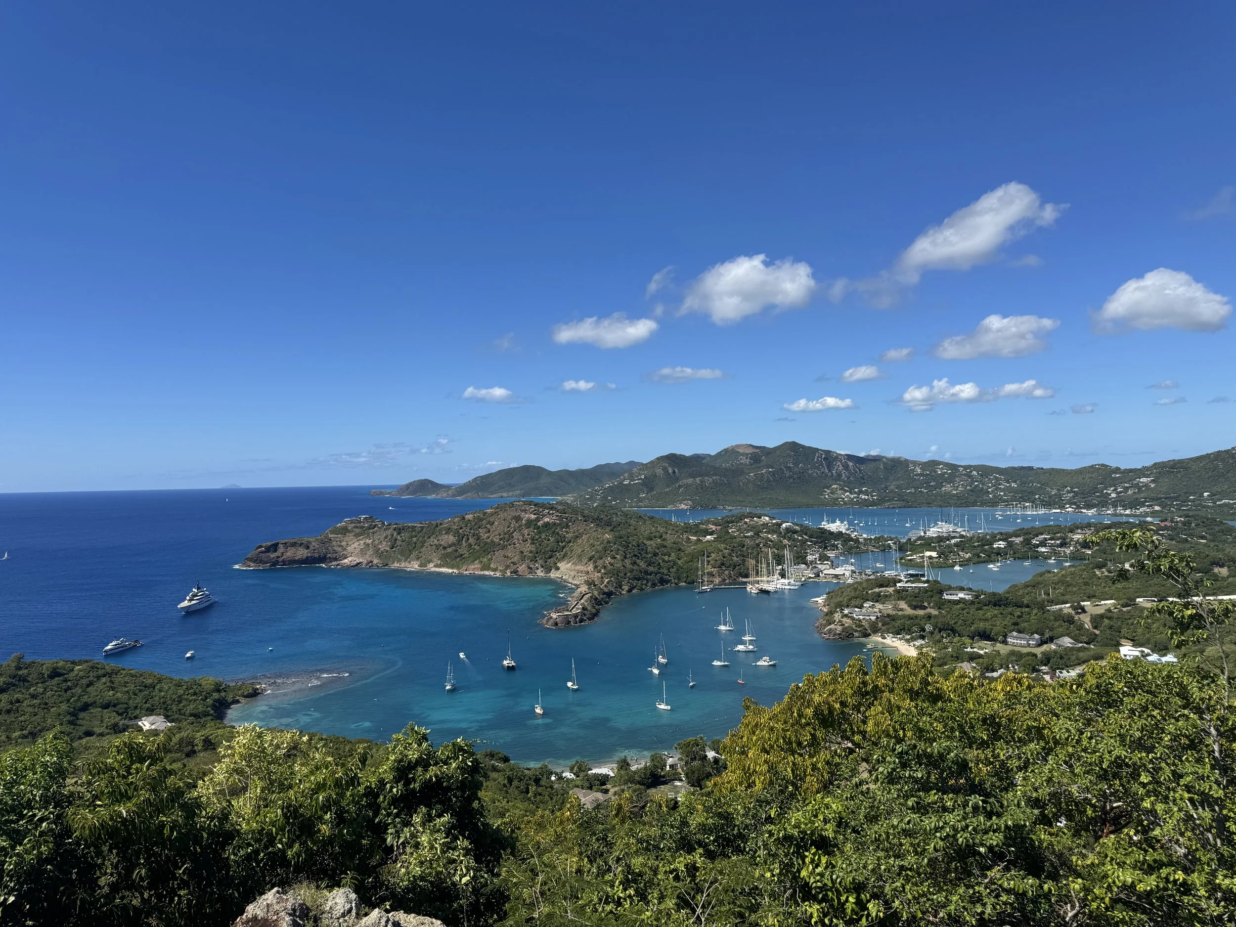 View of a tropical bay with sailboats and yachts, surrounded by lush green hills under a bright blue sky with scattered white clouds.