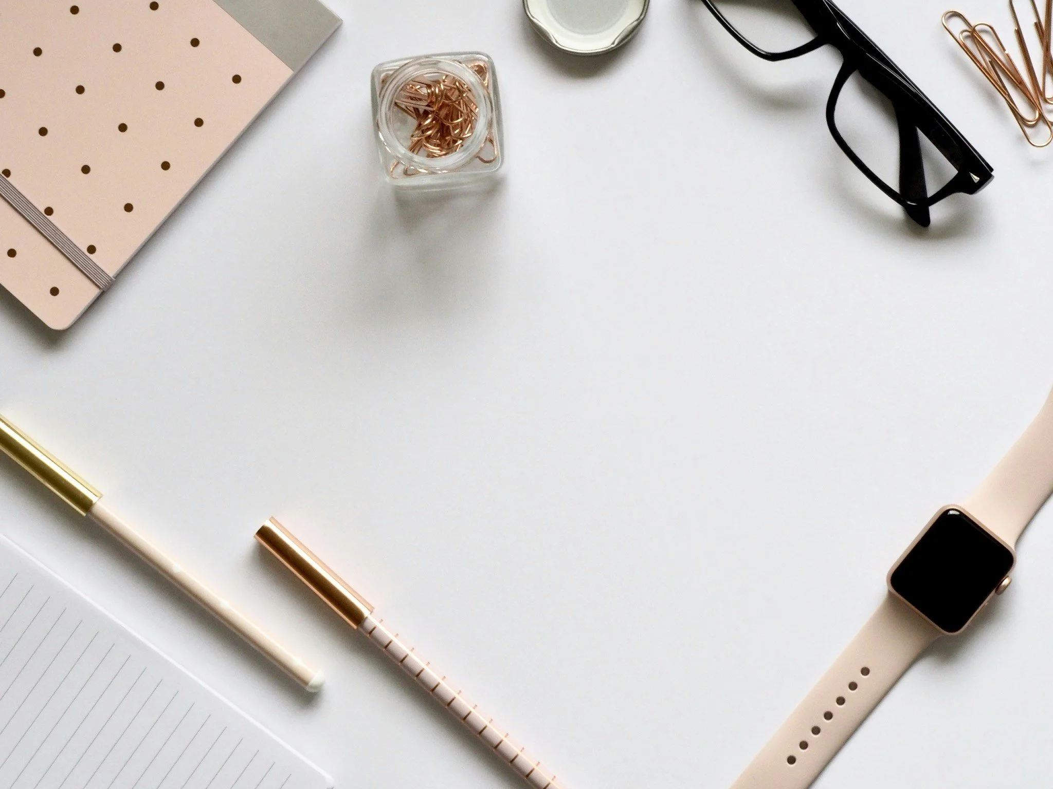 Flat lay of a white desk with a pink polka dot notebook, a white lined notepad, a gold and white pen, a pink and gold striped pen, a small glass container with gold paper clips, black glasses, a white smart watch, and a black device with a white strap.