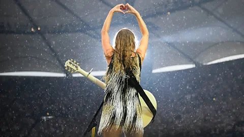 Taylor Swift standing on stage with her back to the audience, arms raised in a heart shape above her head while holding a guitar during the Eras Tour concert.
