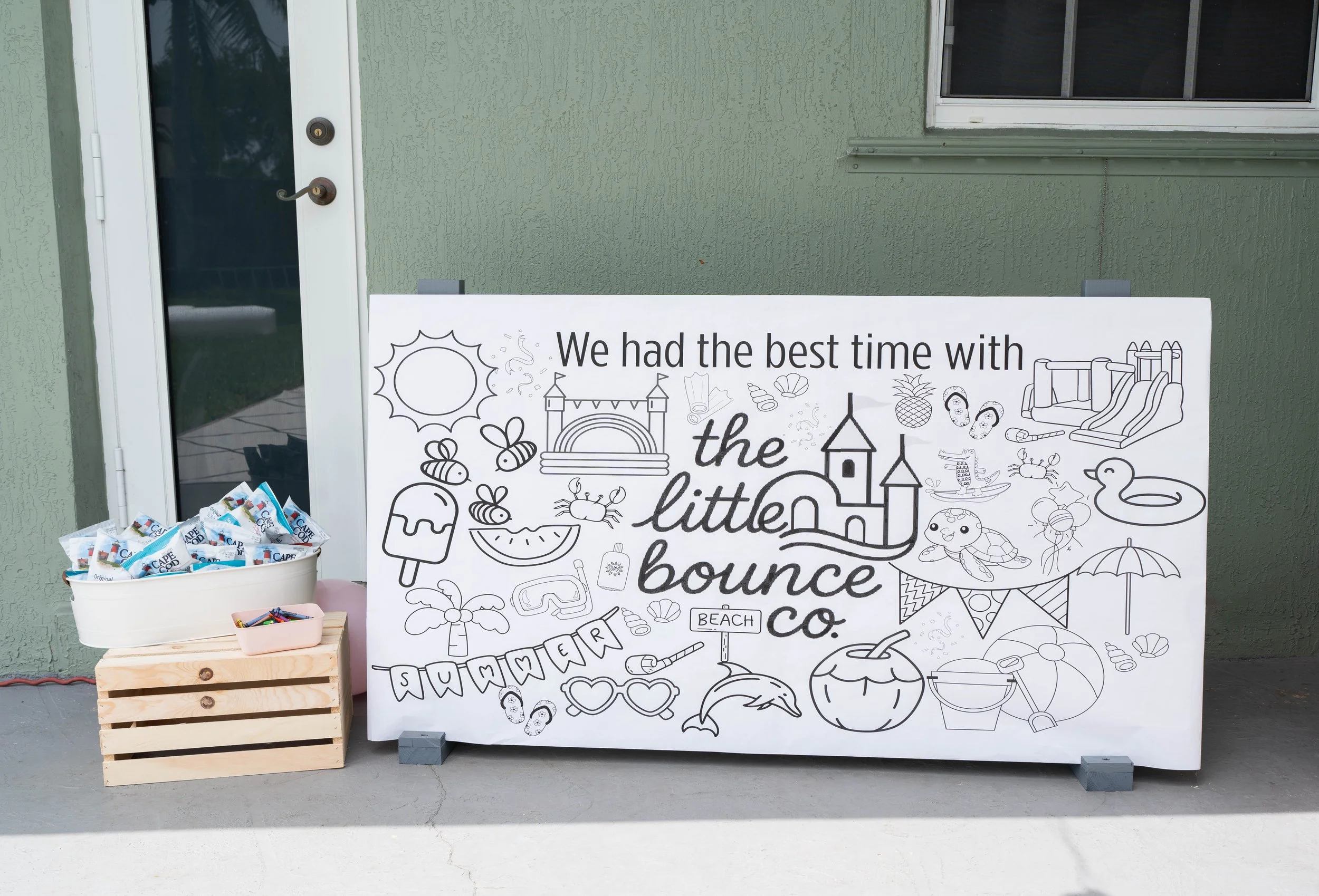 A large white sign with black line drawings and the words 'We had the best time with the little bounce co.' The drawings include a sun, bees, a castle, a mermaid tail, sunglasses, a watermelon slice, beach items, and a slide. Two wooden crates and a white container with snack wrappers and small items are placed beside the sign against a green wall with a window and door.