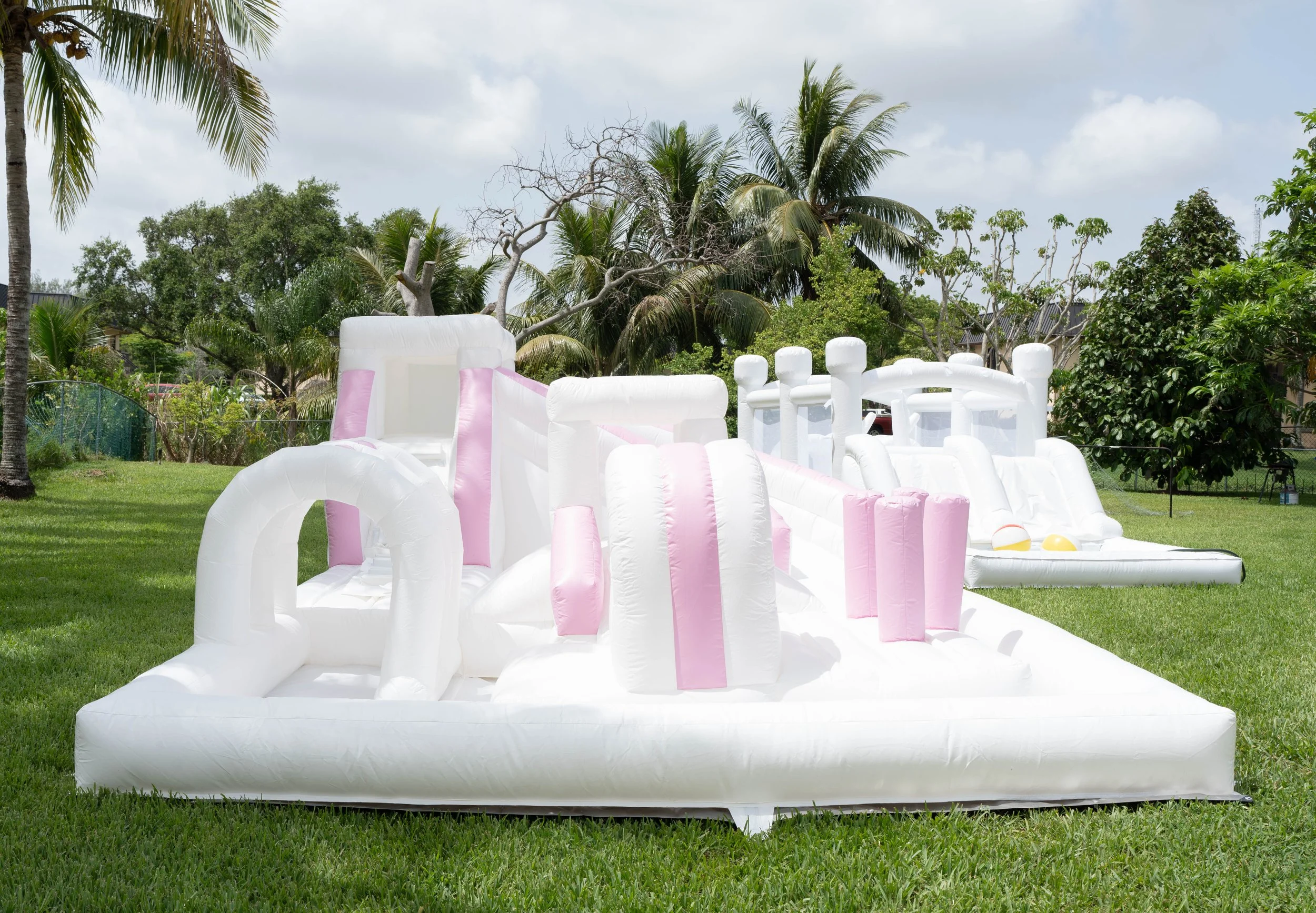 A large white and pink inflatable bounce house designed as a castle with turrets, towers, and slides, set up on a grassy lawn with trees and a partly cloudy sky in the background.