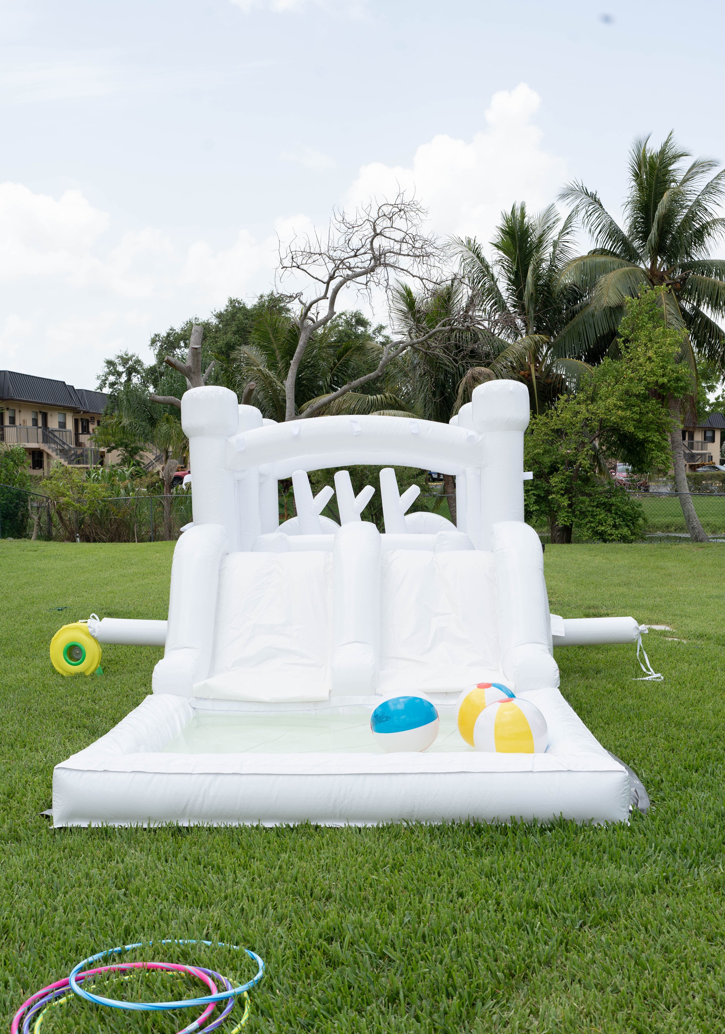 White inflatable water slide with a pool at the bottom, placed on a grassy lawn, with several beach balls and hula hoops nearby, and a backdrop of trees and apartment buildings under a partly cloudy sky.