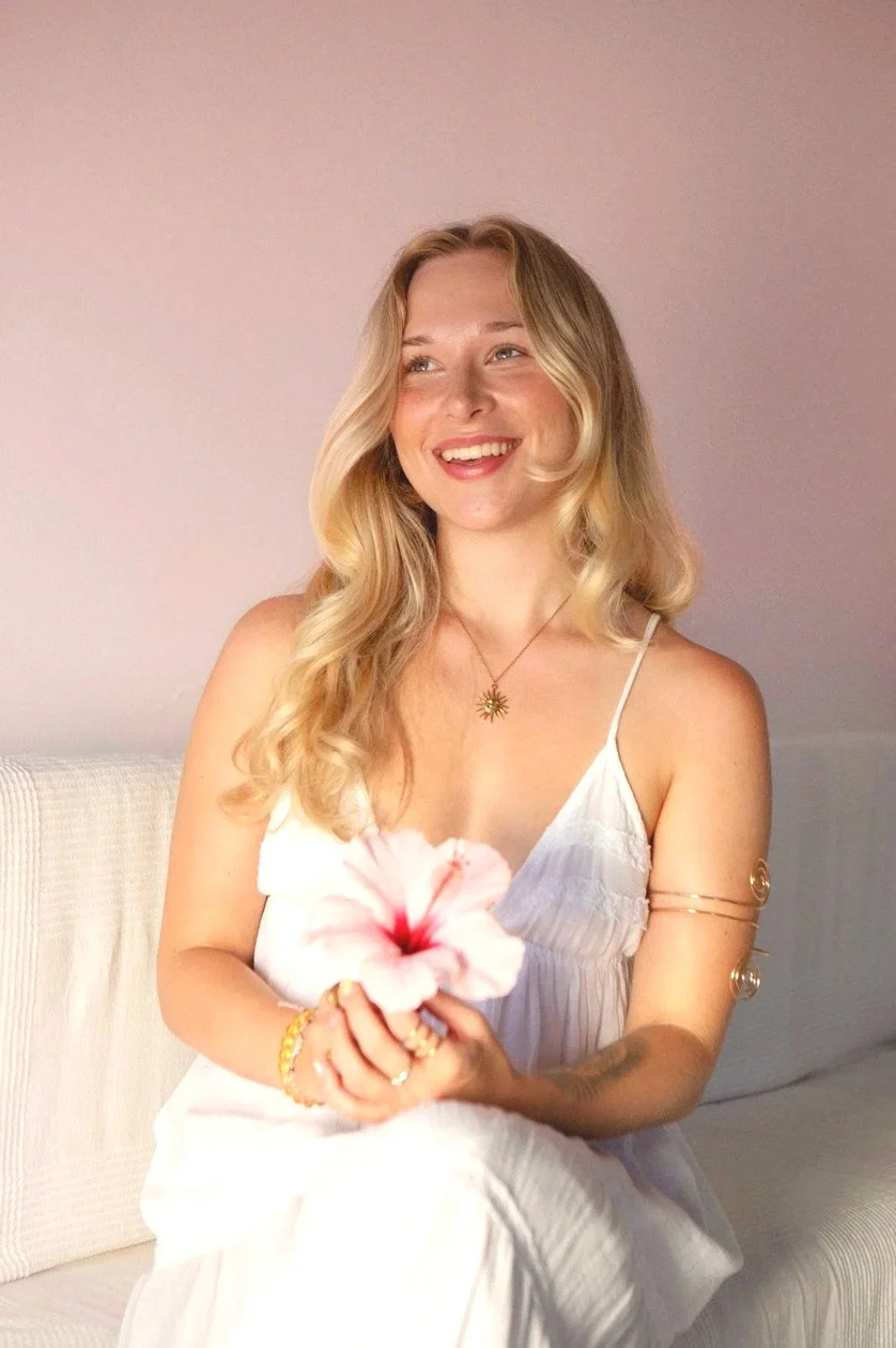 A young woman with long blonde hair, smiling, wearing a white sleeveless dress, holding a pink hibiscus flower, sitting on a light-colored couch with a plain wall background.