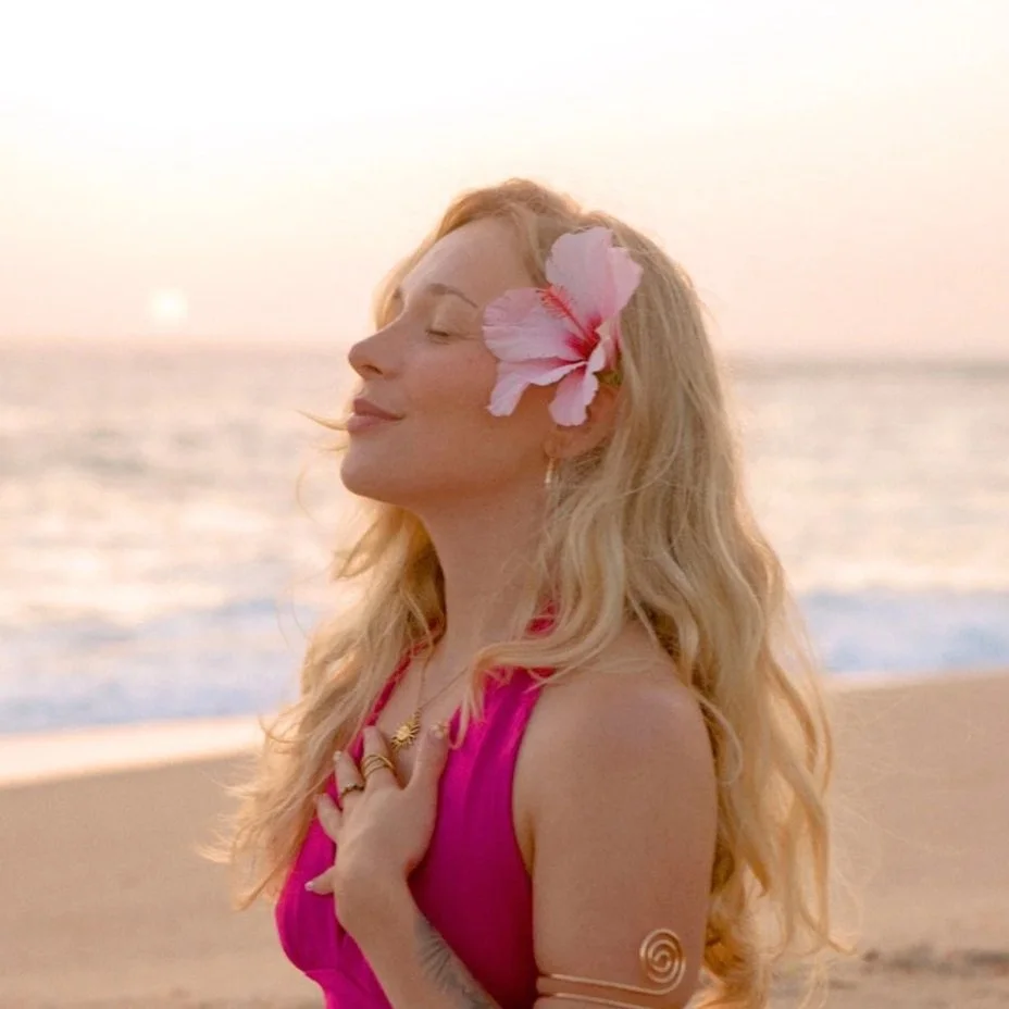A woman with long blonde hair, wearing a pink dress, closed eyes, and a pink flower in her hair, stands on the beach at sunset, with her hand on her chest.