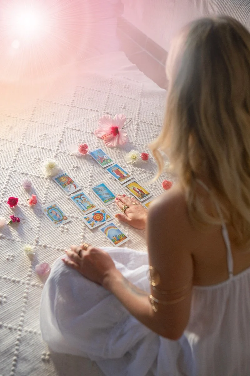 A woman sitting on a white textured blanket with tarot cards laid out in front of her. The scene is decorated with large pink and white flowers, small flowers, and petals. Soft sunlight is shining from the top left corner.