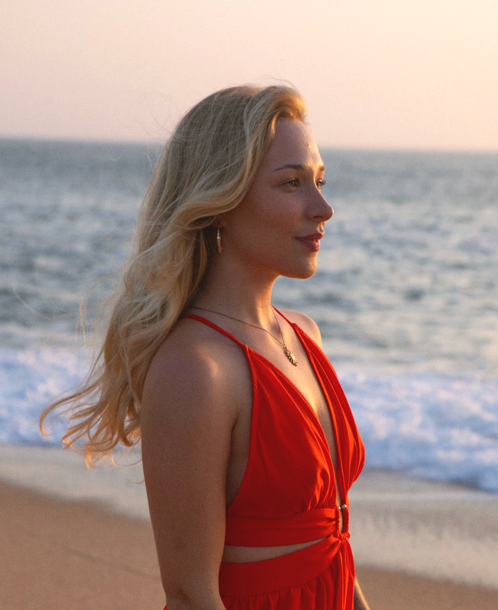 A woman with long blonde hair standing on a beach during sunset, wearing a red dress and jewelry, looking towards the ocean.