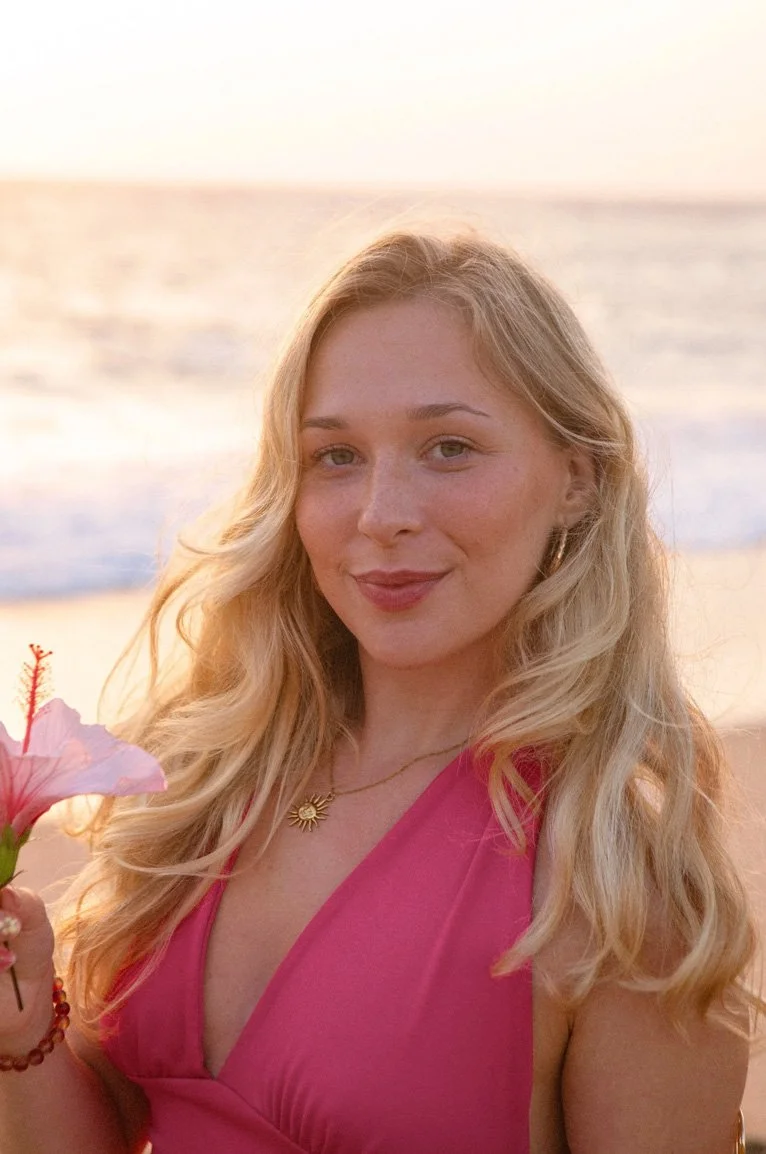 A young woman with long blonde hair smiling at the camera, holding a pink hibiscus flower, with the beach and ocean in the background during sunset.
