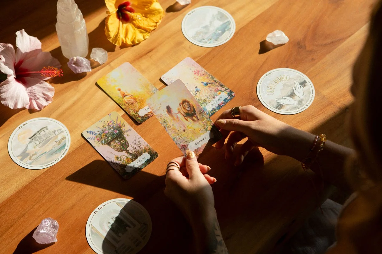 A person holding tarot cards at a wooden table, surrounded by flowers, crystals, and circular tarot spread mats.