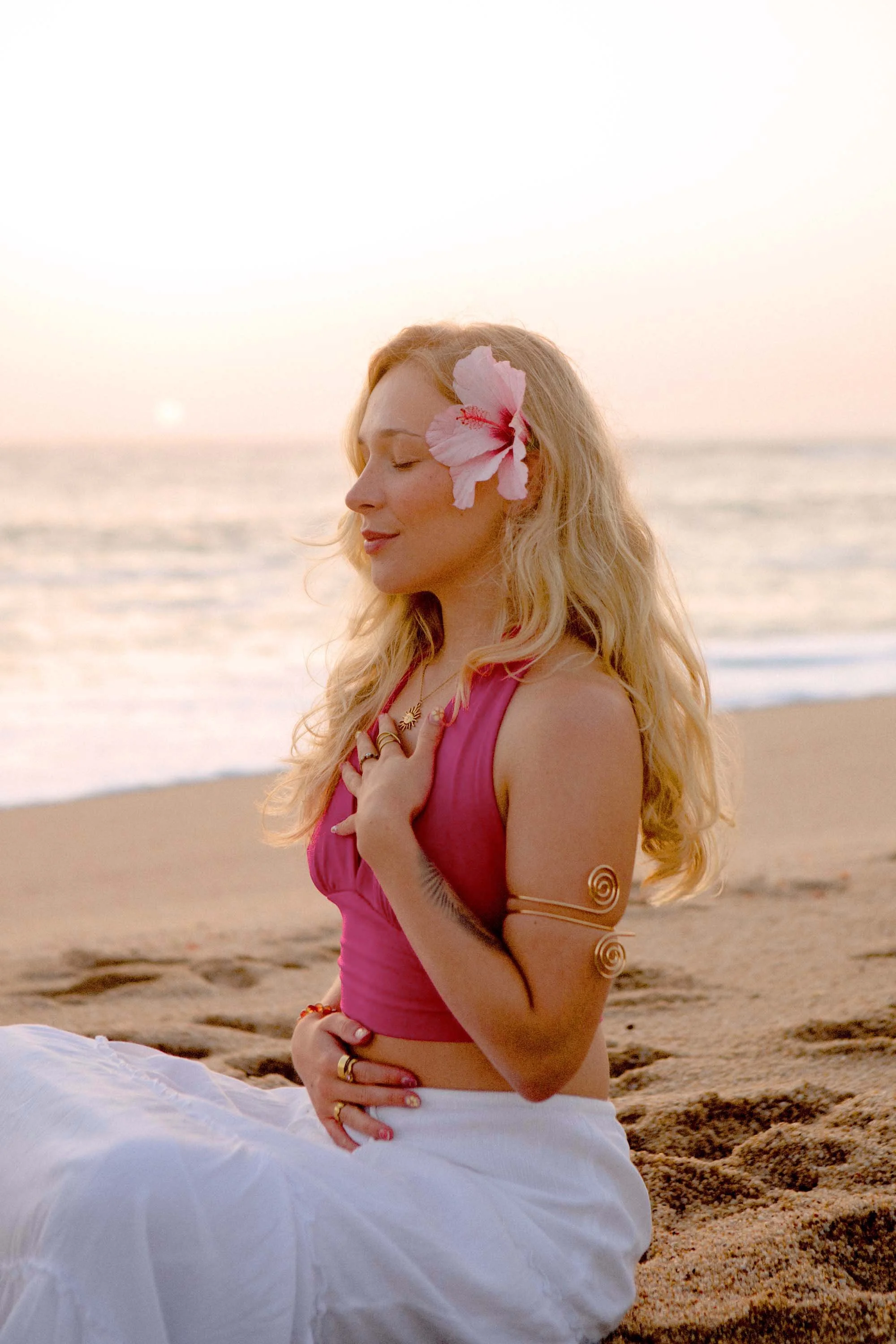 A woman with long blonde hair sitting on the beach during sunset, with a pink flower in her hair, wearing a pink top, white skirt, and jewelry, with her eyes closed and hands crossed over her chest.