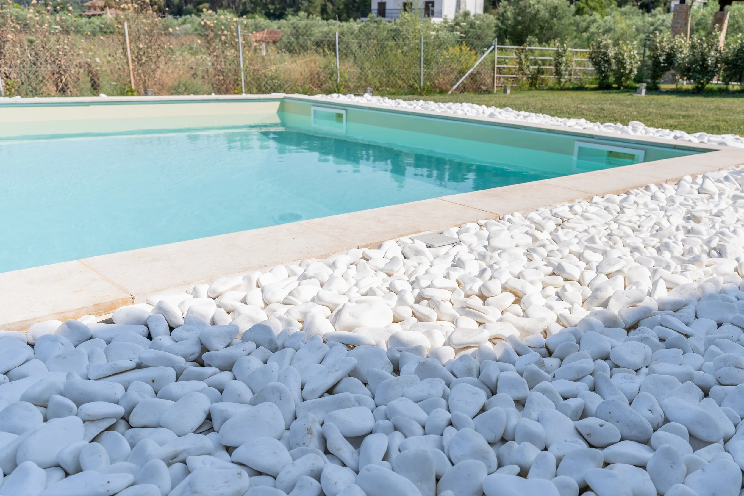 A backyard swimming pool with white decorative rocks surrounding it and a grassy area with trees and bushes in the background.