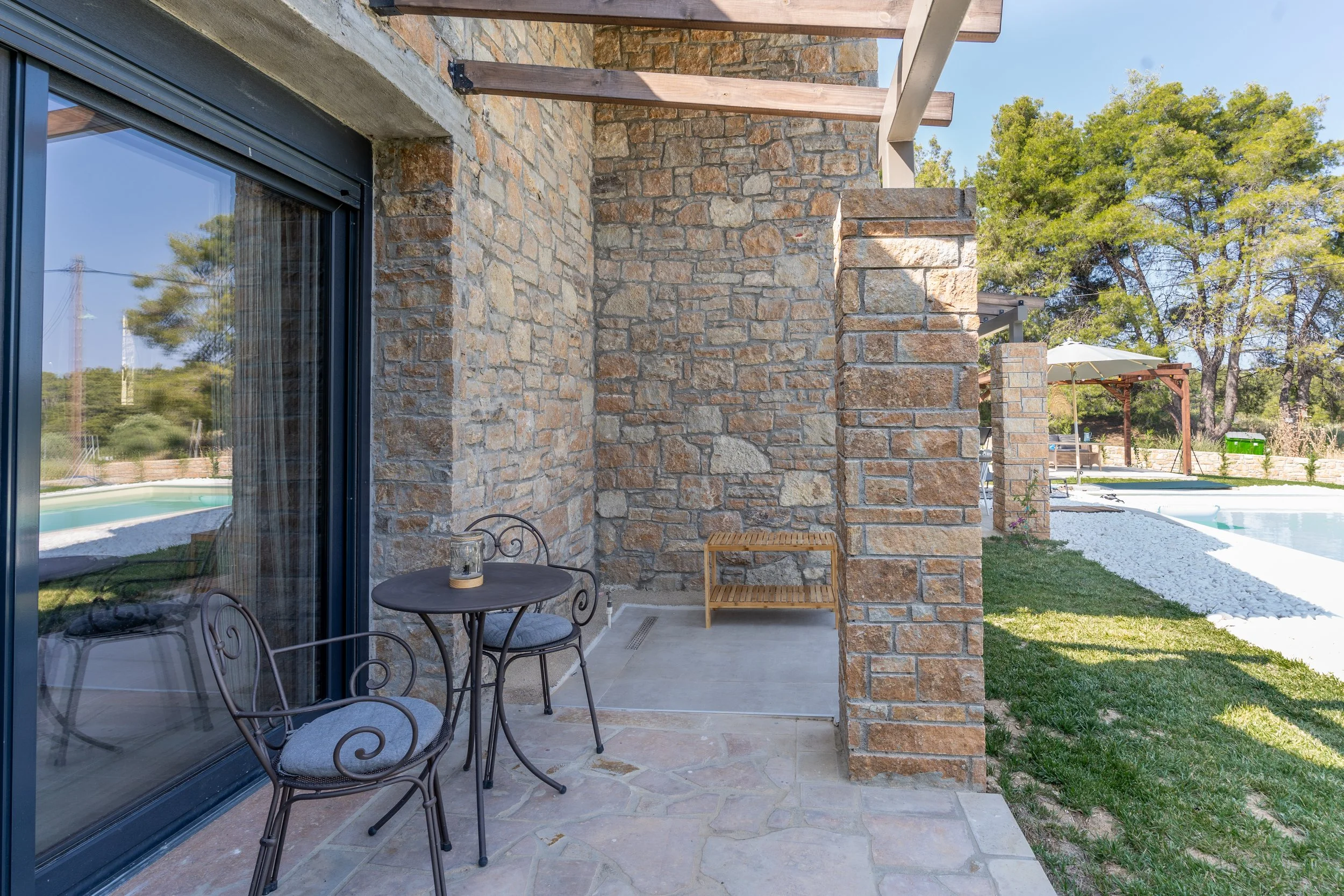 Outdoor patio area with Metal table and two chairs, brick wall, sliding glass door reflecting pool, grassy yard, canopy-covered seating area, trees, and clear blue sky in background.