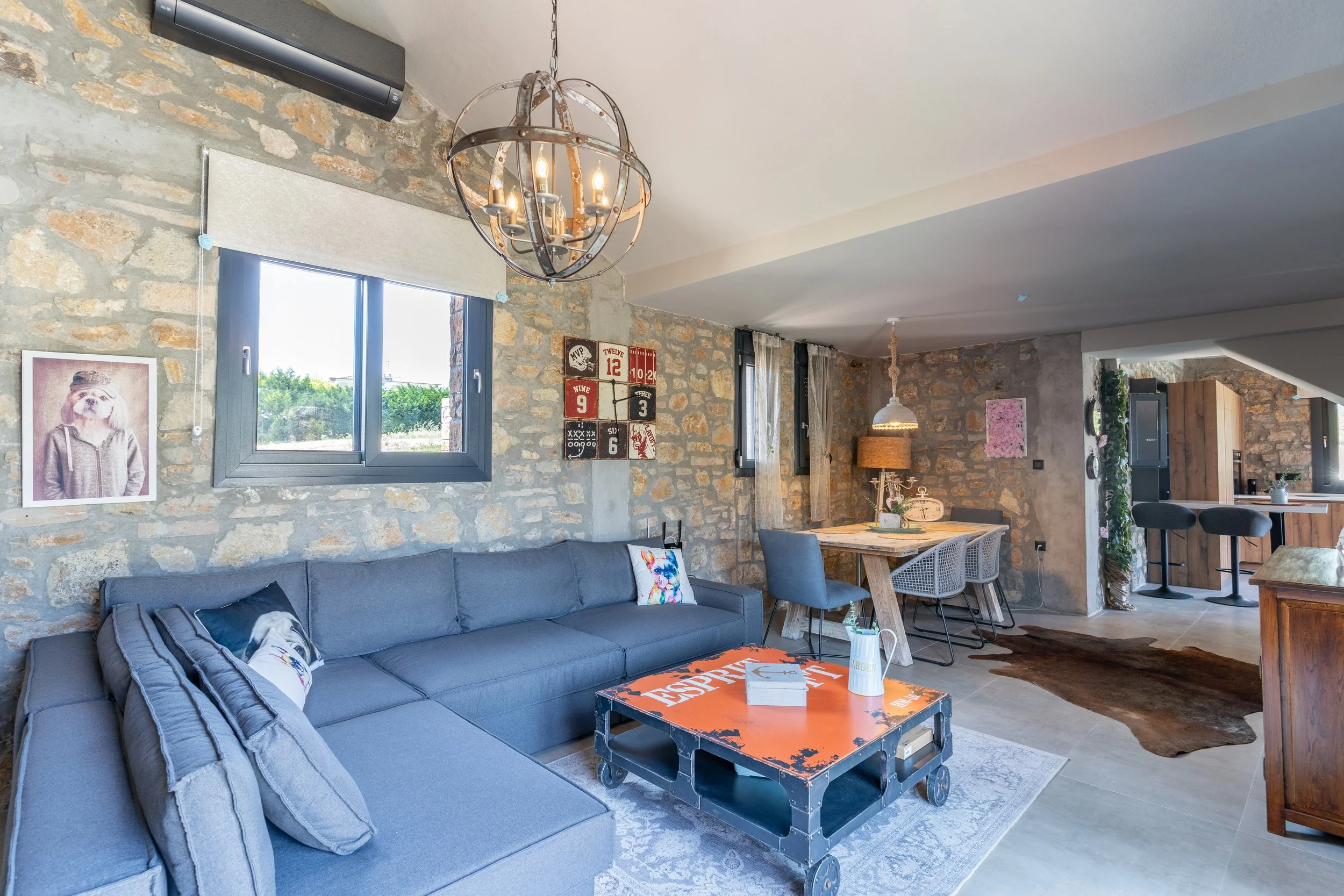 Living room with exposed brick wall, gray sectional sofa, colorful pillows, wooden coffee table, wall art, and dining area with table and chairs, modern light fixtures, and a view of the kitchen.
